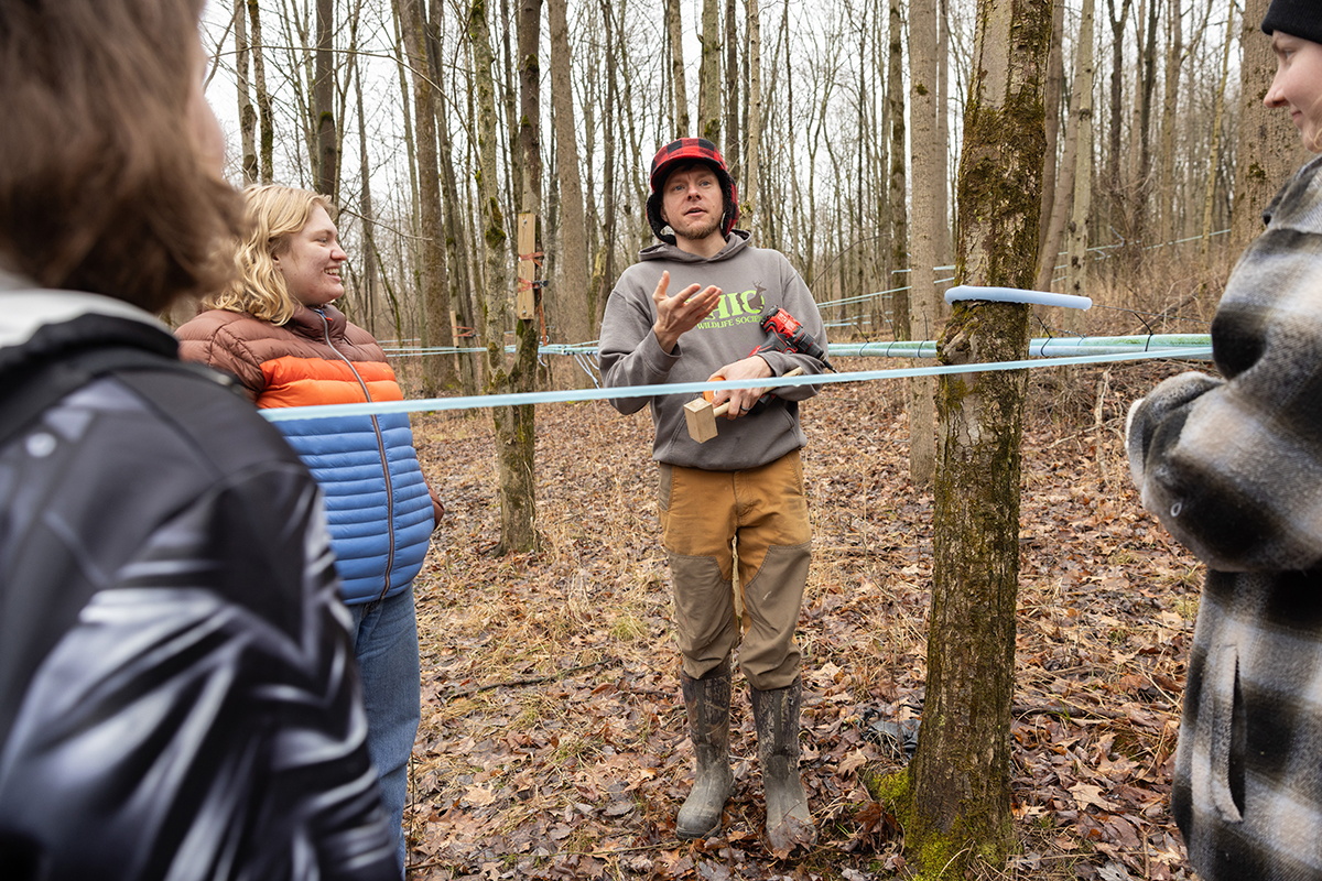 Gabe Karns, a thin white man wearing a plaid hunter's cap, gestures as he talks to students in the woods. Blue and green flexible rubber tubes, about an inch or two thick, weave through the forest around them. The student, three females dressed for cool temperatures, smile as they listen.