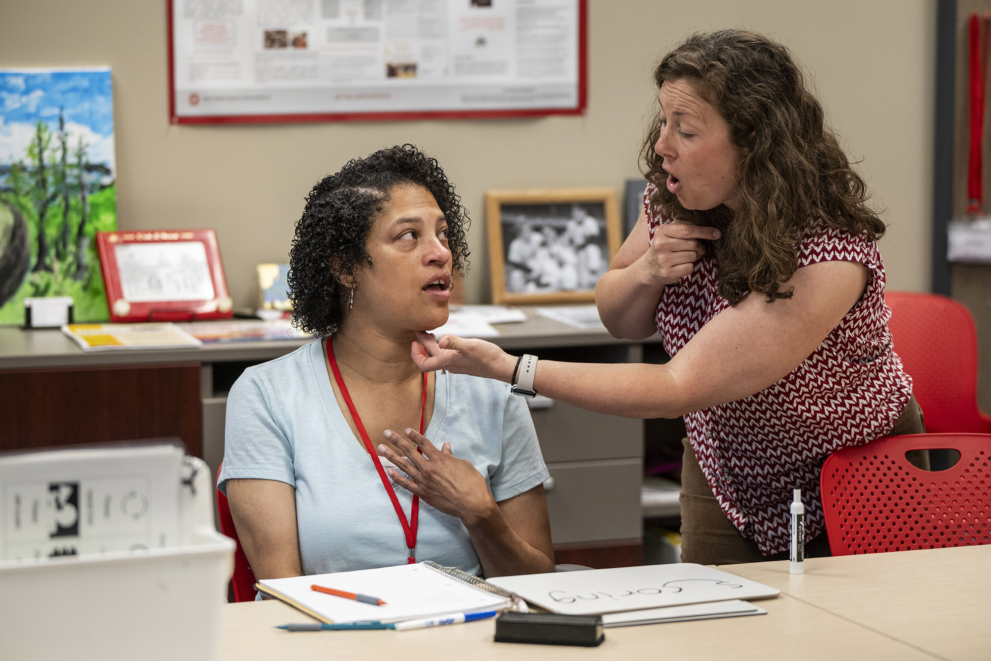 Amber Poindexter, sitting at a table, touches her chest as she watches the mouth of her speech therapist, Arin Sheeler. Arin is a curly-haired white woman leaning toward Amber, pointing at her own throat and touching the underside of Amber’s chin. They’re very focused on each other.