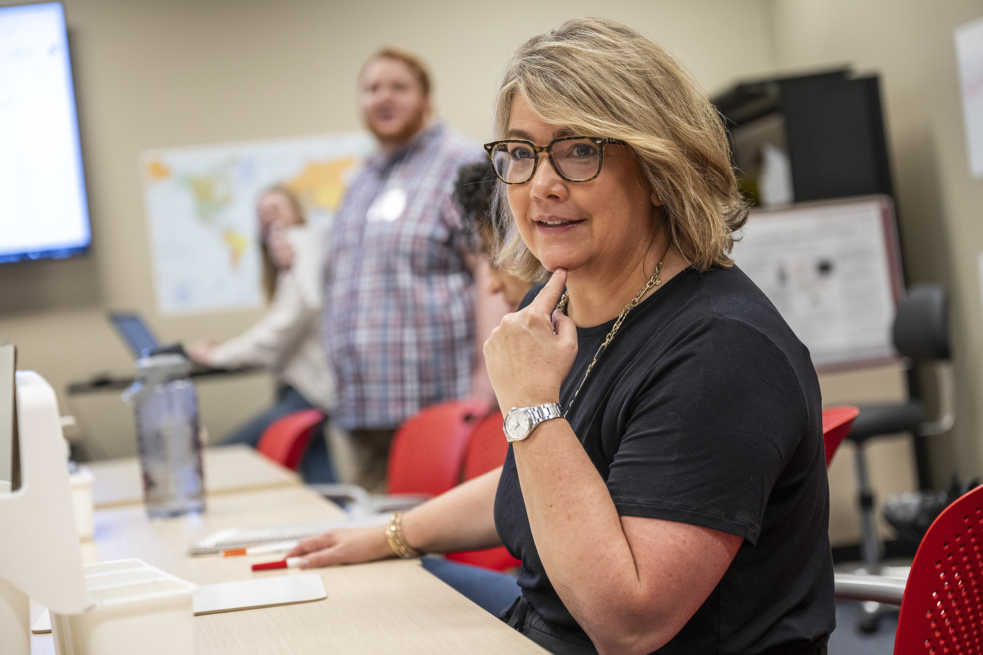 Jennifer Brello, a middle-aged white woman, points at her chin as she watches someone outside of the photo frame. Out of focus behind her are people who participate in the Aphasia Initiative programming. She has blond, tussled hair that’s just past chin length and wears glasses, a long necklace and a silver watch.