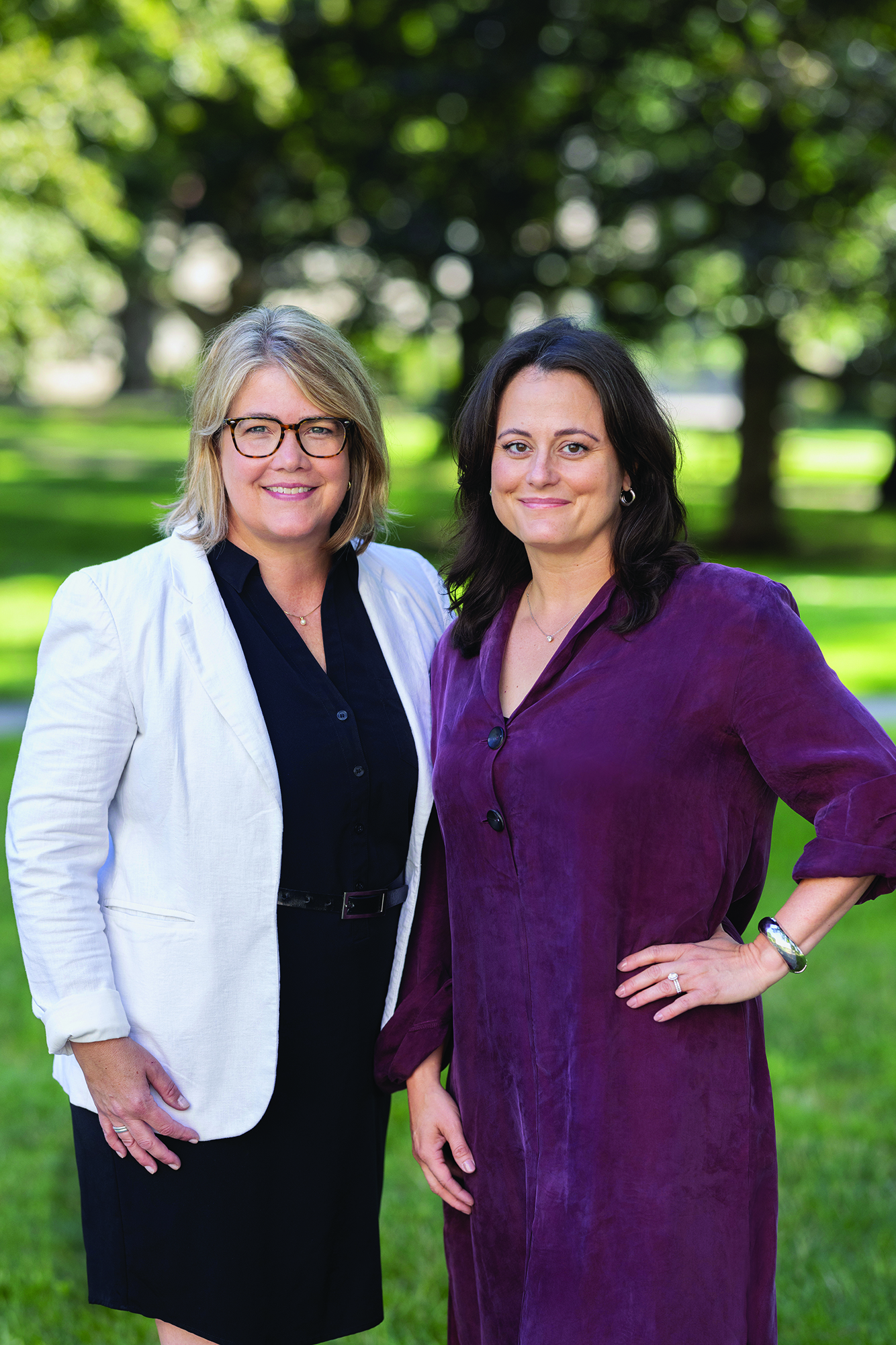 Posing on Ohio State’s Oval, which is out of focus behind them, are Jennifer Brello and Stacy Harnish. Both are attractive middle-age white women who are smiling. Jennifer is blonde and wears glasses and a sport jacket over a tailored dress. Stacy is a brunette wearing a shift-style dress and stands with a hand on her hip. They are close, seemingly comfortable with each other.