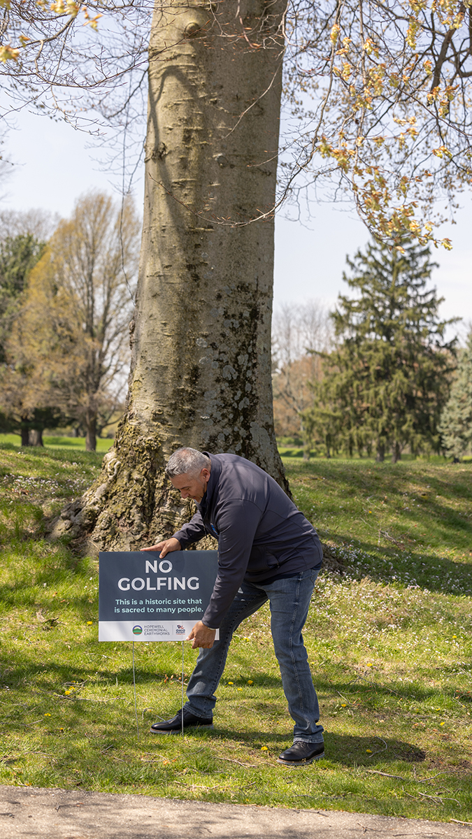 A man pokes a yard sign into the grass in front of a massive tree. The sign says "No golfing," a reference to when the earthworks site was owned by a golf course.