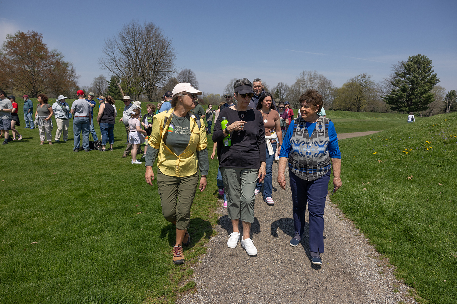 Three women talk as they walk with a crowd through the Octagon Earthworks on a sunny day.