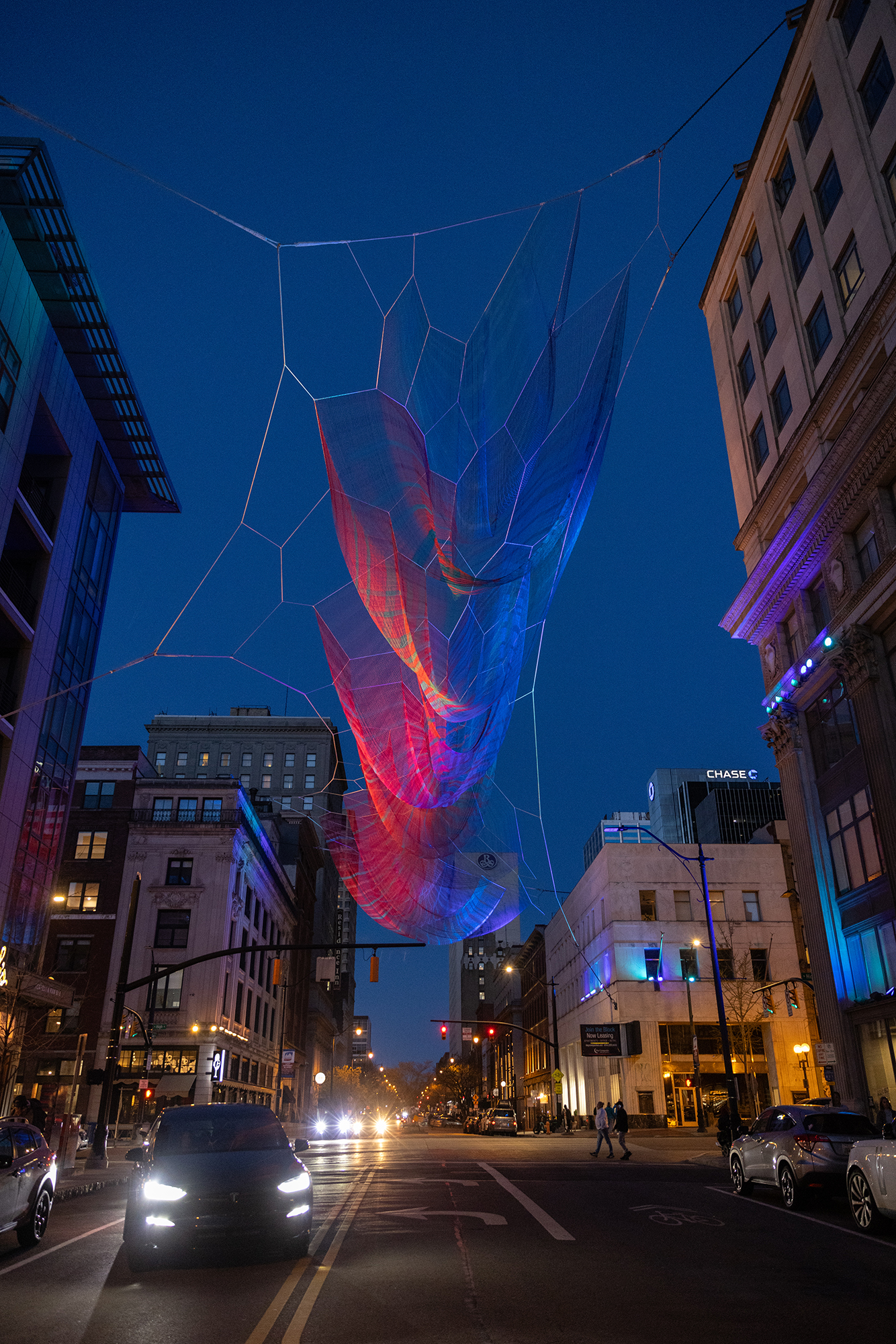 A sculpture suspended between buildings across from each other on High Street in Downtown Columbus. The wires form different geometric shapes and transparent fabric swoops down from those shapes.