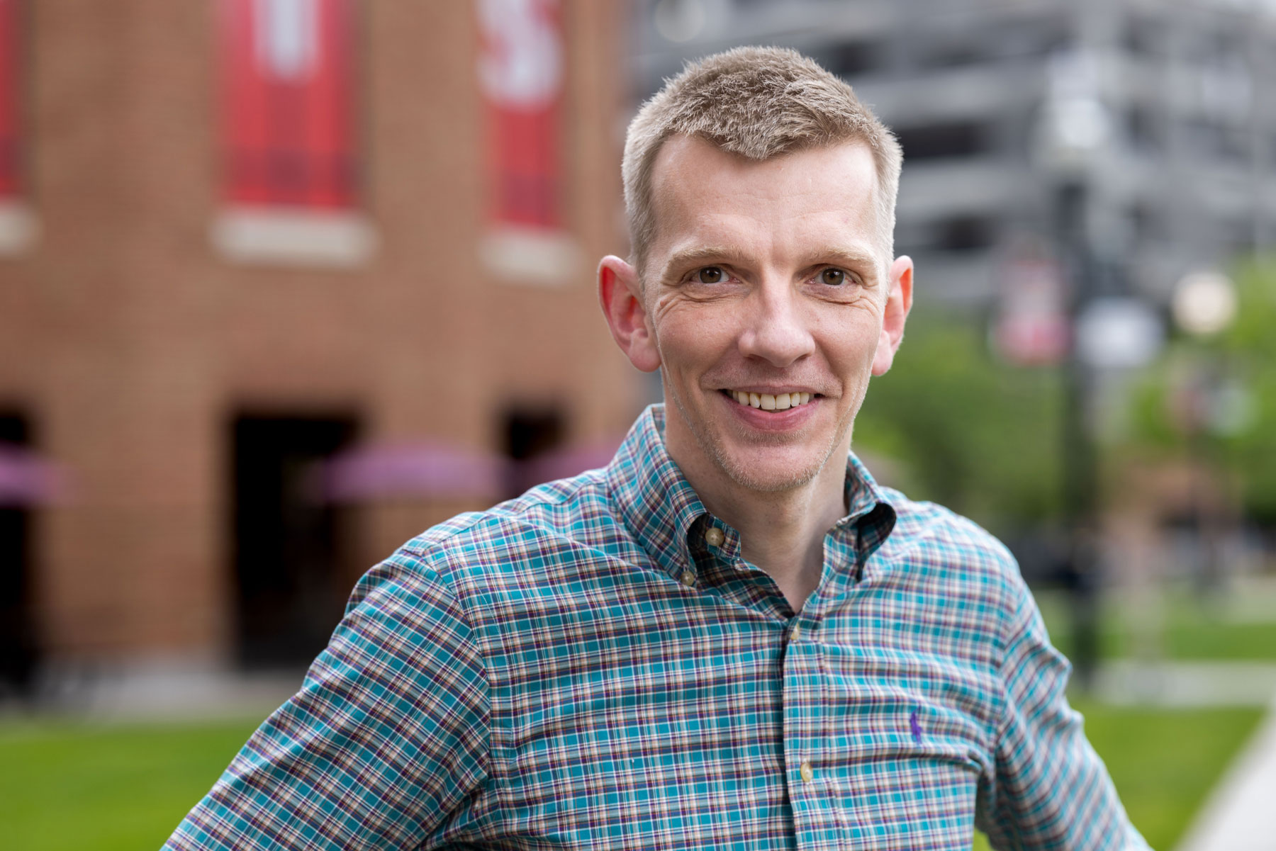 Angus Fletcher, a white man with neatly trimmed short hair, smiles and wears a patterned button-up shirt while standing outdoors with hands on hips. A brick building and a campus background with greenery appear behind him.