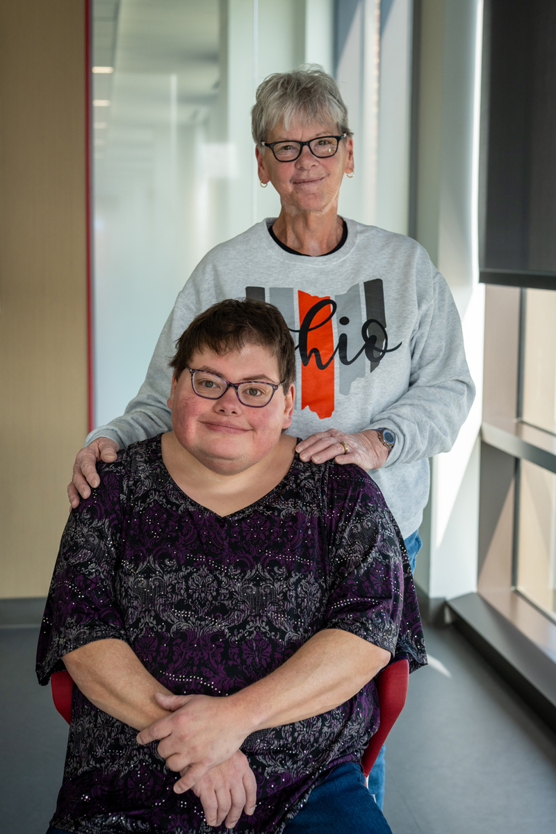 A mother stands behind her adult daughter.
