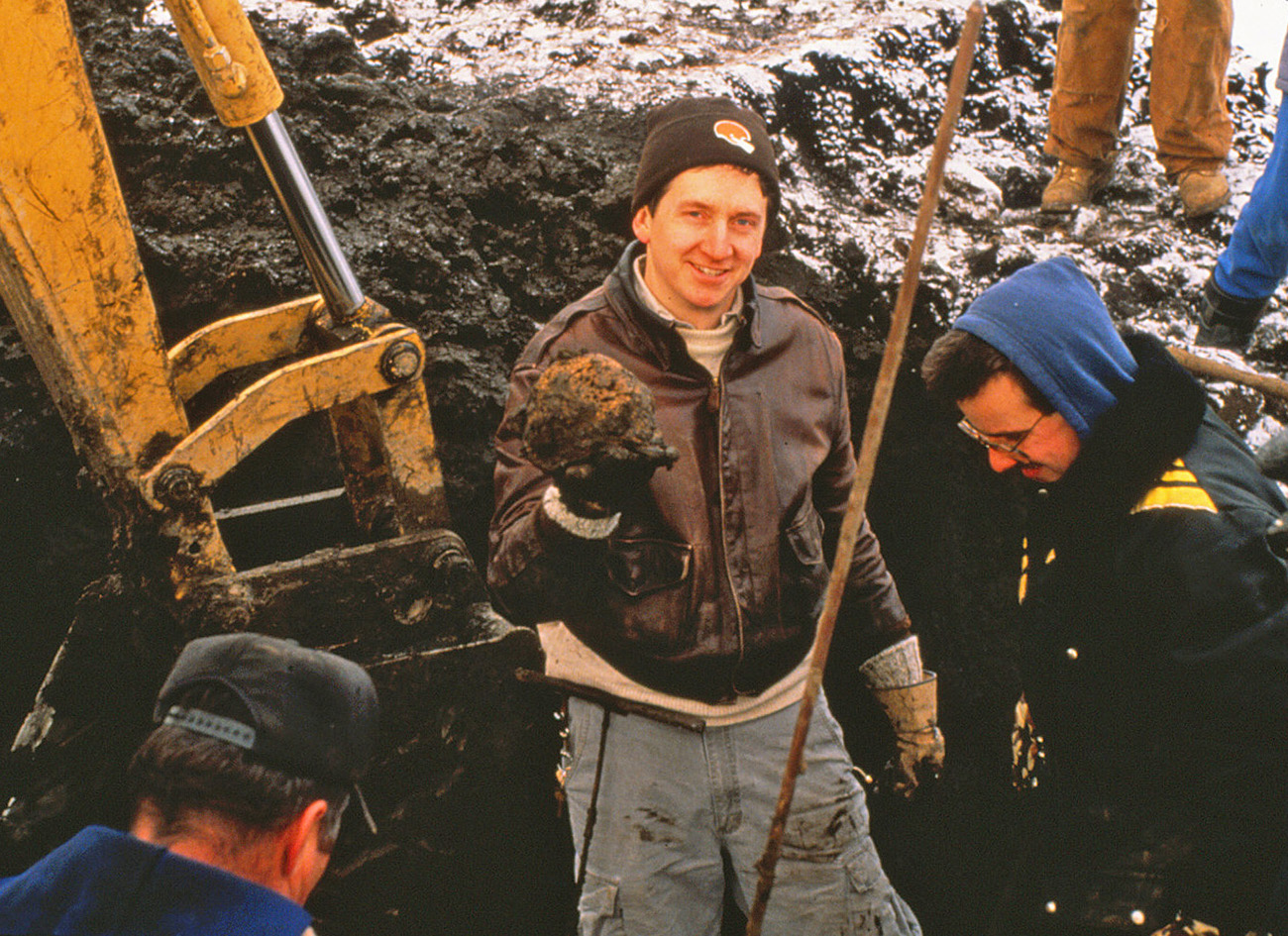 A younger Bradley Lepper, dressed in a leather bomber jacket and Cleveland Browns stocking hat, stands in a dirt pit surrounded by other people surveying or digging. He smiles as he shows a muddy round thing, smaller than a basketball but bigger than a softball. It is a part of a mastodon skeleton.
