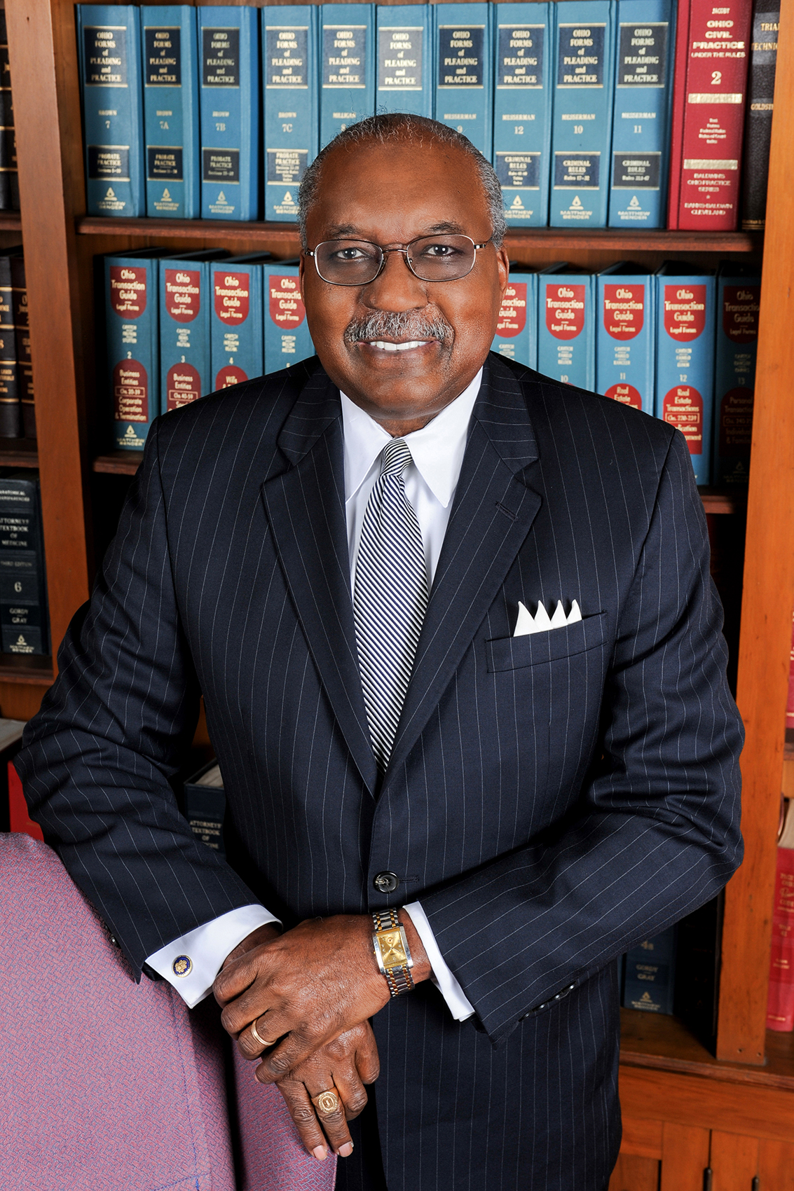 This portrait of Ben Espy shows a sophisticated Black man with a friendly smile. He wears glasses and a pin-striped suit and stands in front of shelves lined with law books.