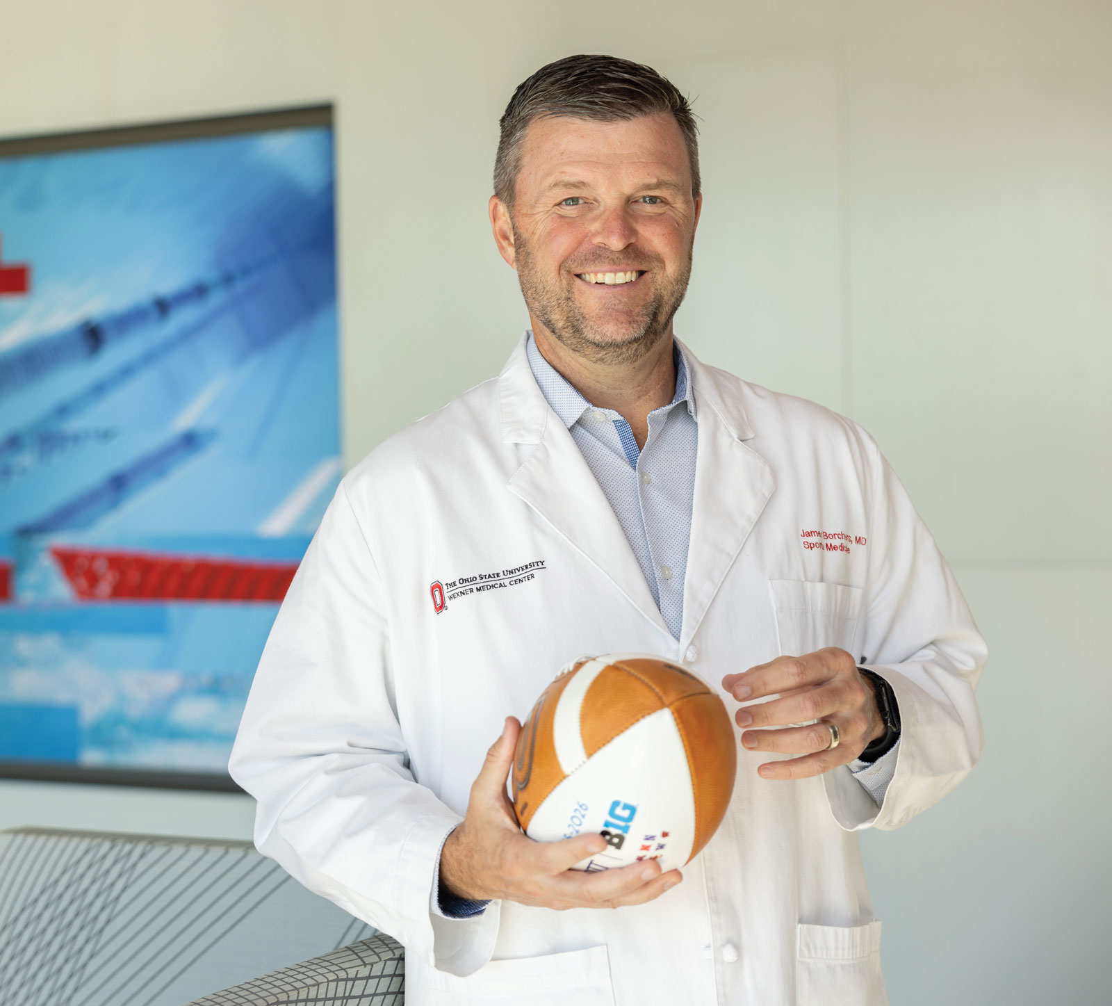 A doctor wearing a medical coat holds a football and smiles as he has his portrait taken.