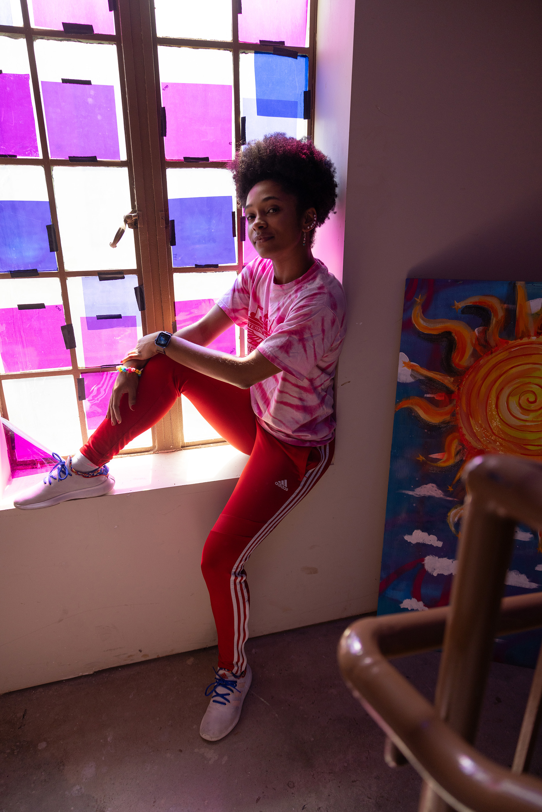 Brrianna Rhodes, a pretty, young black woman, sits on a window ledge inside a dim room. Colored paper taped over the window shines colored light onto her. She's smiling with her eyes and wearing casual clothes that she can dance in. Her hair is tight curls on top of her head and she has a line of earrings up the ear the photo shows. 