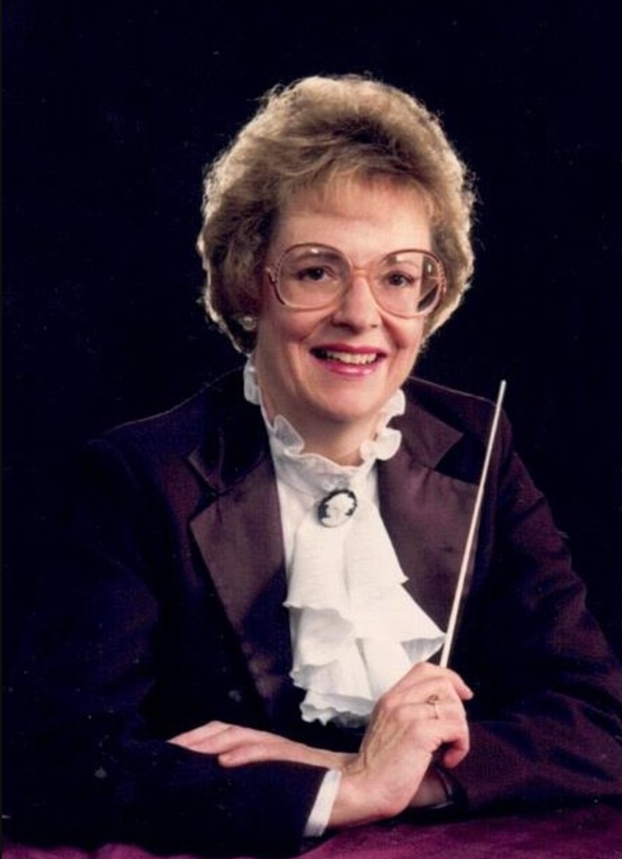 Carol Clark poses for a portrait holding a conductor's baton and dressed for an orchestra. She's smiling, has 1980s-style rounded frame glasses, pearl earrings and short blonde hair attractively poofed.