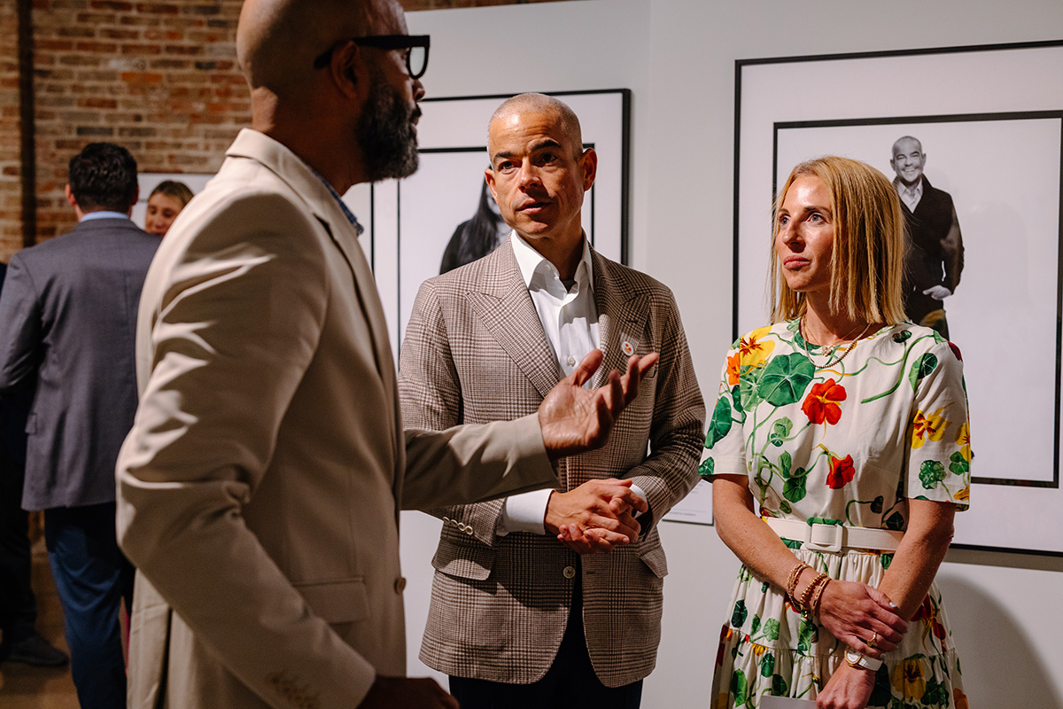 Matt Scantland and his wife listen to a tall man, a photographer, at a gallery exhibit. They're ll dressed up and seem to be having an interesting conversation.