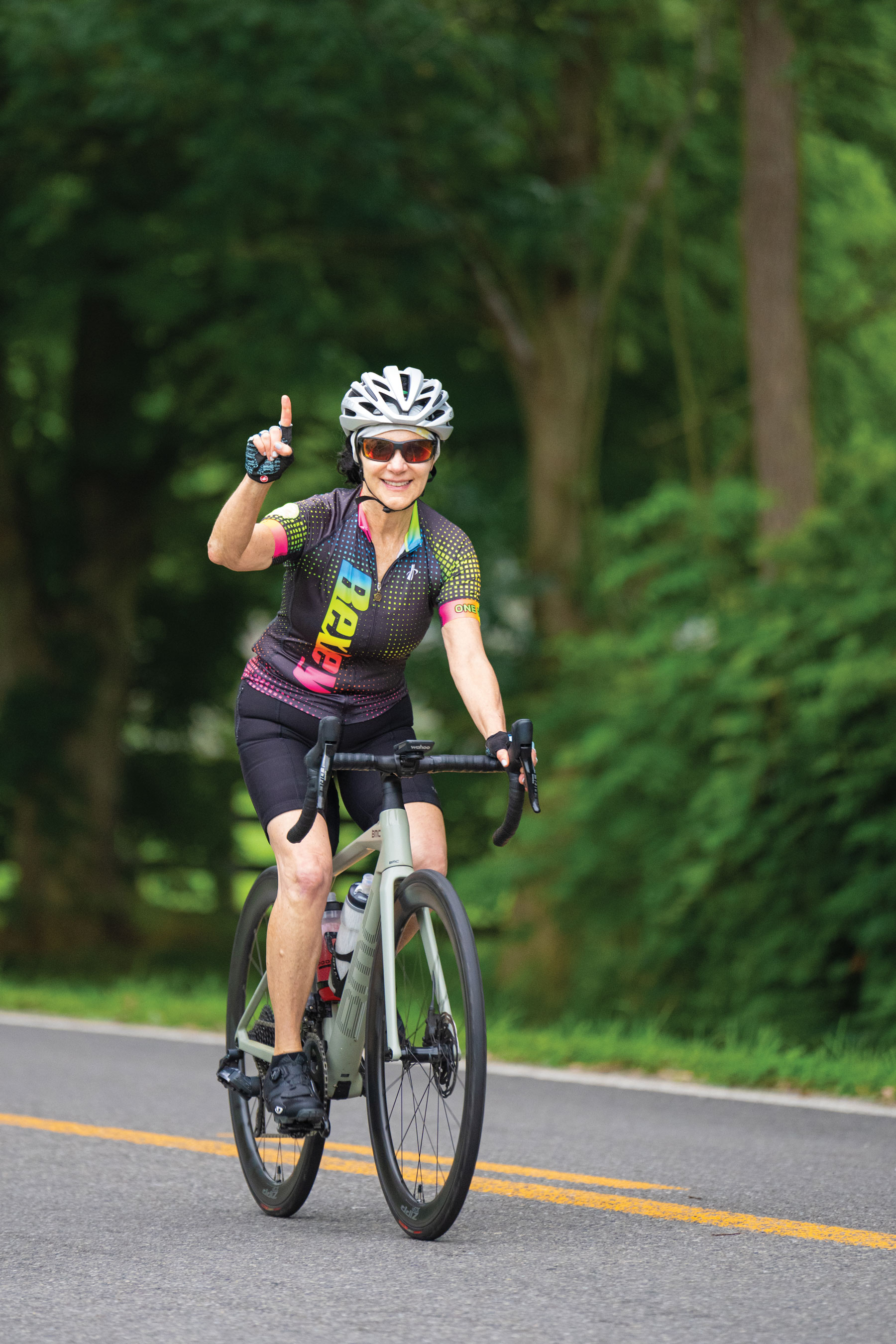 riding a bicycle down a tree-lined street, Linda Kass raises her finger in the number one symbol. She wears a helmet and bike jersey that says Bexley.
