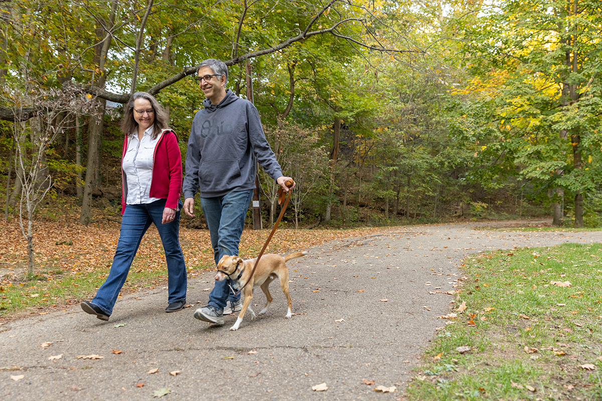 A smiling couple walks their dog through woods on a paved path