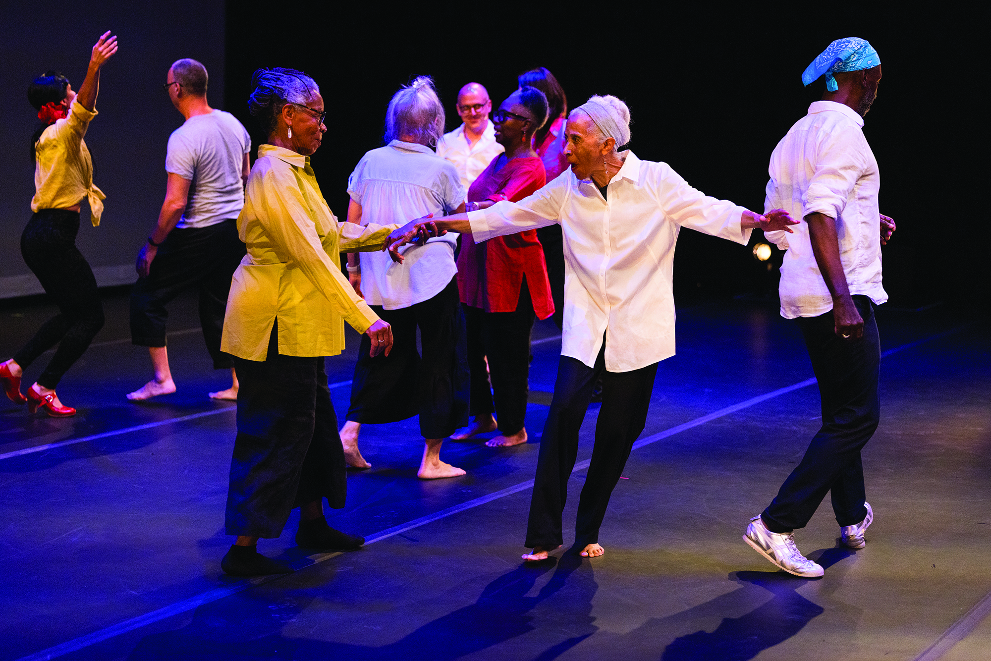 Amid dancers dancing on stage, Dianne McIntyre greets Bebe Miller. Both women wear loose black pants and big button-down shirts. Dianne's expression shows how pleased she is to dance with Bebe; she has a big smile as they make eye contact. Bebe's face, shown from the side, seems to be welcoming back an old friend. Both women are black; the other dancers are a mix of races.