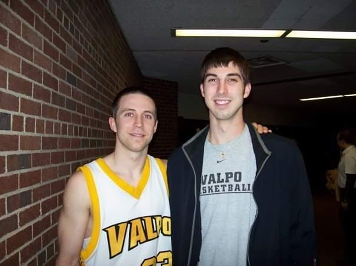 In their college days, Jake Diebler and his brother, Jon, pose together in a hallway at what looks like a sports facility. Jake wears a basketball jersey and has his arm wrapped around the shoulders of his taller brother, who is in street clothes. Both smile.