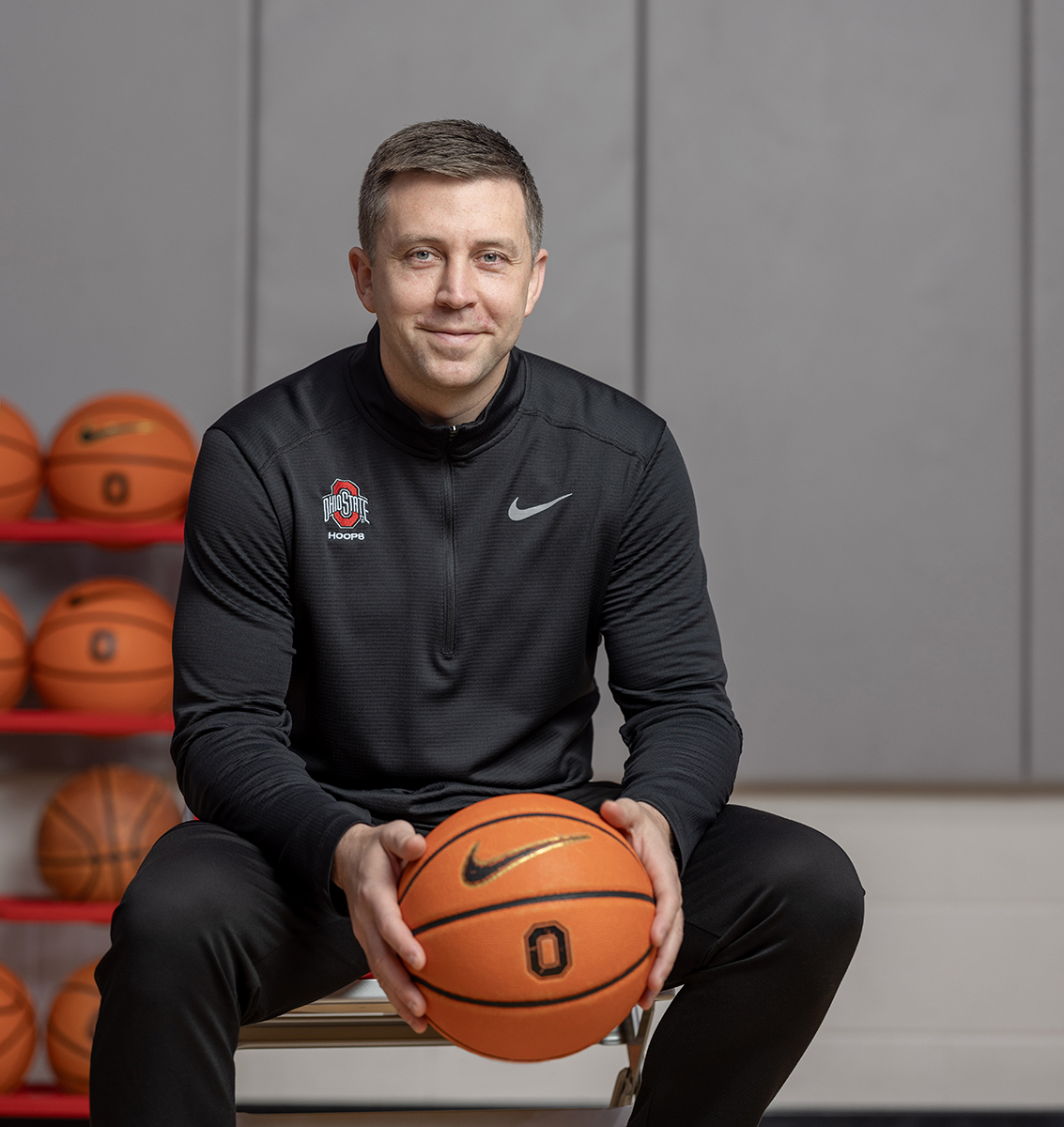 Ohio State Basketball Coach Jake Diebler holds a basketball while sitting on a stool and smiling warmly at the camera. He looks like he is about to laugh. Diebler is a white man who looks to be in his 30s. He has short cropped hair, blue eyes and wears black workout gear.