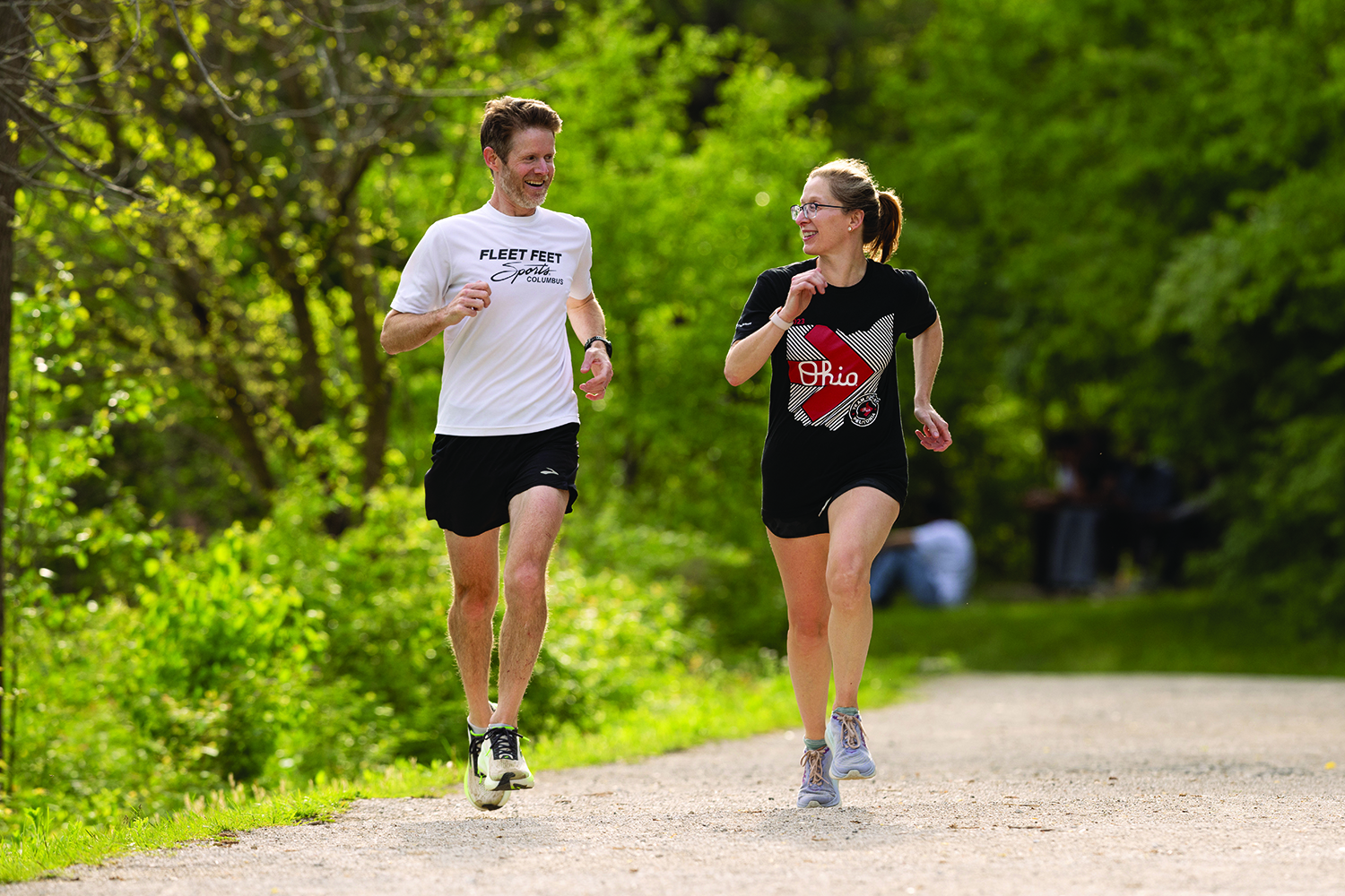 A woman and man talk as they run down a bicycle trail surrounded by trees and lush vegetation.