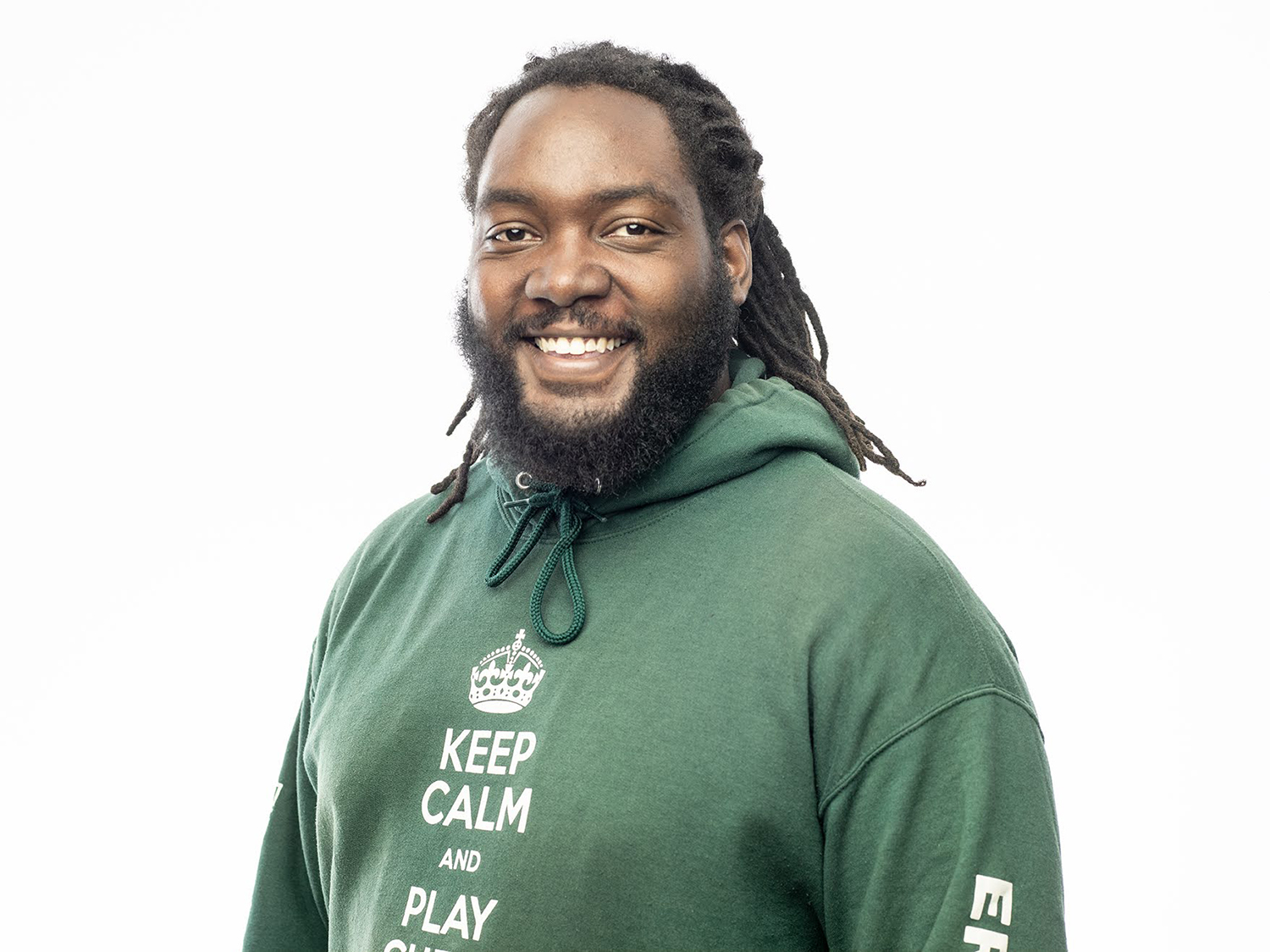 In this portrait of Ernest Levert Jr., he poses with a friendly smile. His sweatshort says "Keep calm and play chess" and has a beard and thin dreads pulled back