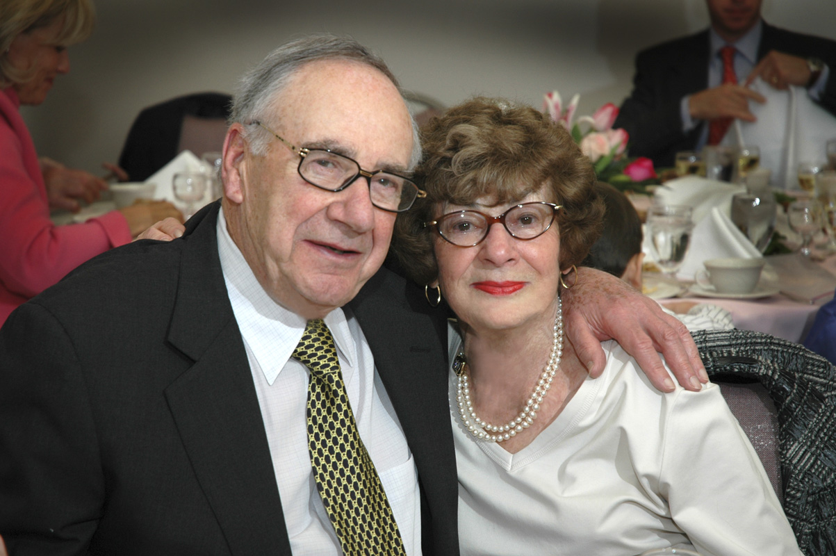 AN older couple proudly pose for a photo at a family event, such as a wedding. They're dressed well and look happy to be sitting with arms around each other.