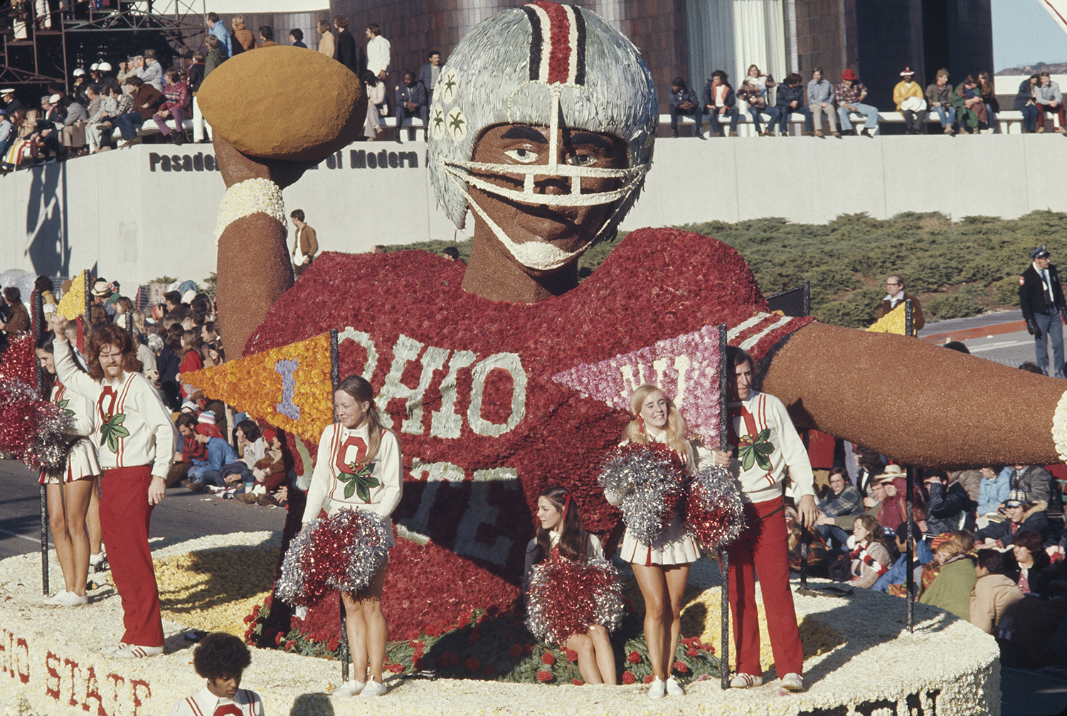 An Ohio State float in the Rose Bowl Parade has a giant football player with arm cocked back to launch the football. The player is black and has a helmet with the signature buckeye leaf stickers that awarded for amazing plays. Standing around him riding the float are male and female cheerleaders. It looks to the the 1960s or '70s, judging by hair and clothing styles.