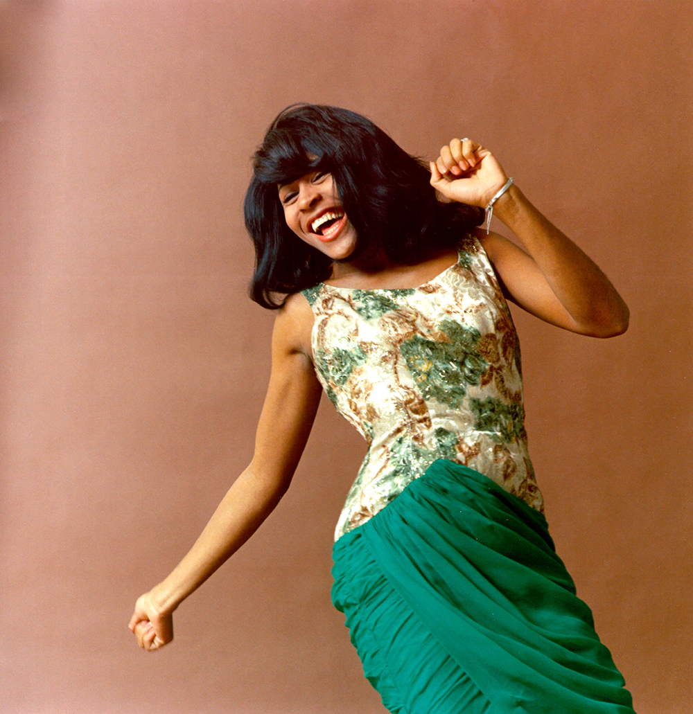 Tina Turner, a black woman in a pretty dress with flowing shoulder length hair, dances in front of a photo backdrop. She has a big smile and her body shape is reminiscent of a twist-style dance. She seems to be having a lot of fun.