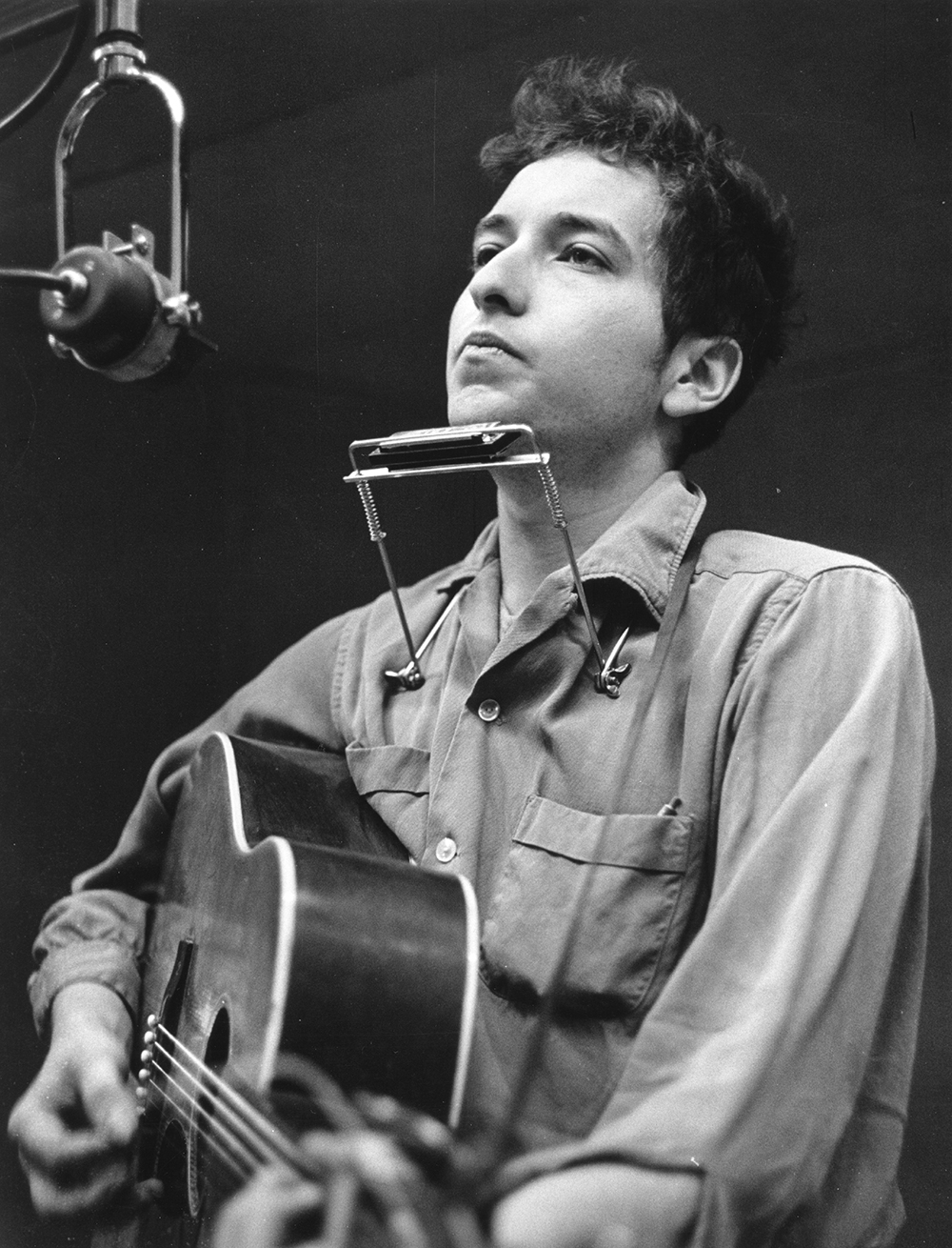 A youn Bob Dylan looks into the distance in a recording studio. A mic is less than a foot from his face and he has his harmonica on a stand hanging around his neck, while he holds his guitar. He looks thoughtful as he perhaps listens to someone not in the photo.
