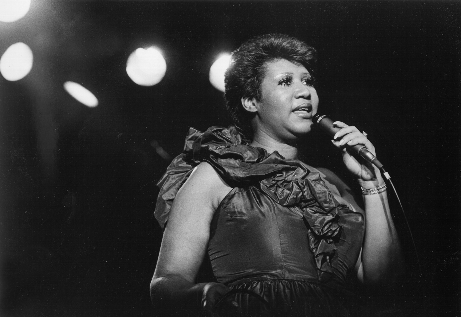 Aretha Franklin, wearing a fancy taffeta-seeming dress, sings into a mic on stage.