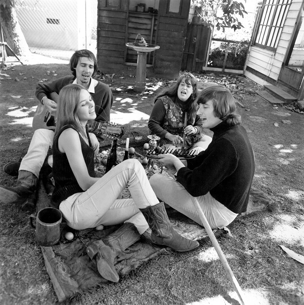 The Mamas & the Papas sit on a dirt ground, seemingly behind someone's house, and sing in a circle. One man plays a guitar. One woman holds a flute. 