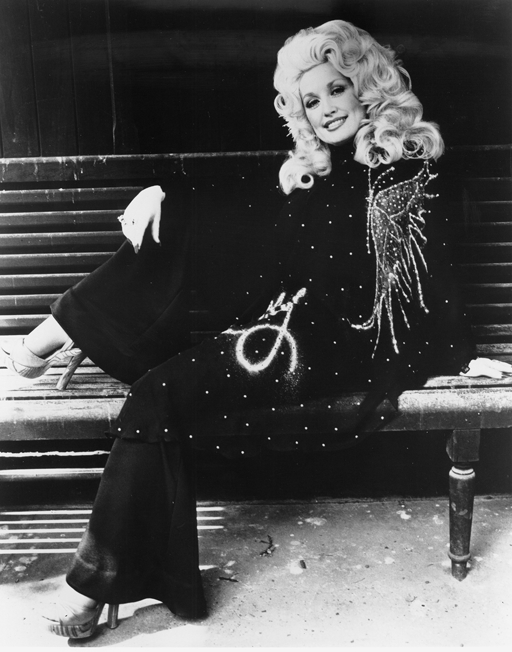 Dolly Parton poses on a bench wearing a dark, drapey pants outfit bedazzled with a rhinestones. She's gorgeous with gorgeous hair, a big smile and eye makeup that is both bold but not that unnatural looking.