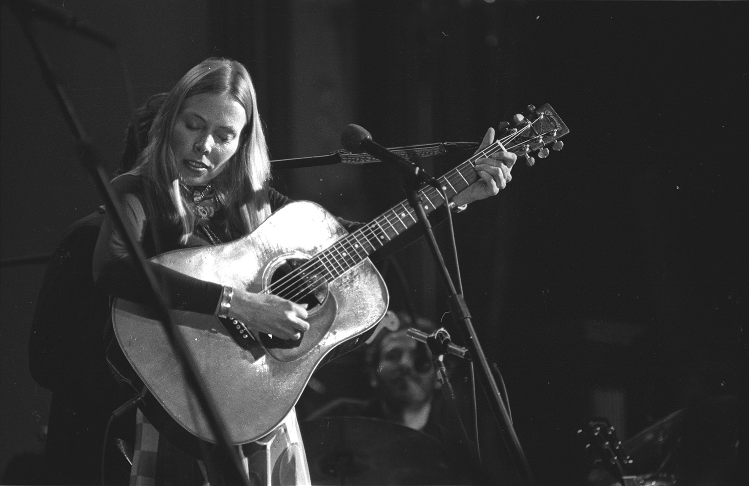 A white woman with straight blond hair, closes her eyes as she holds her guitar out, playing it with emotion. She looks very absorbed by her music.