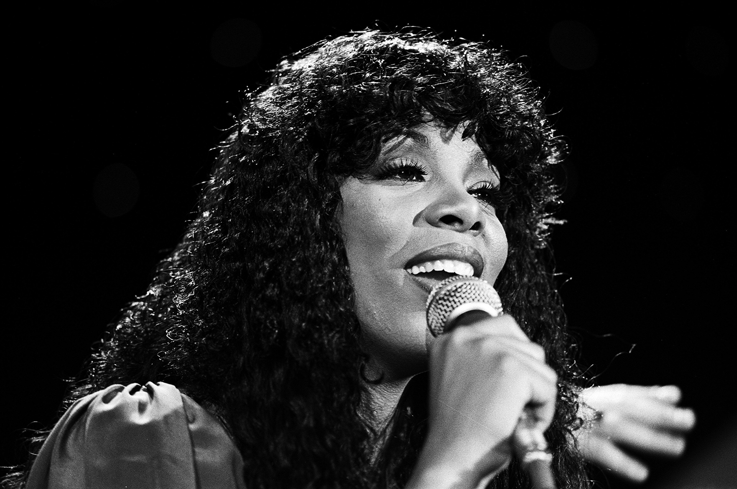 Donna Summer, a pretty, Black woman with long curly hair, sings into a mic while on stage. She's smiling while she does. 