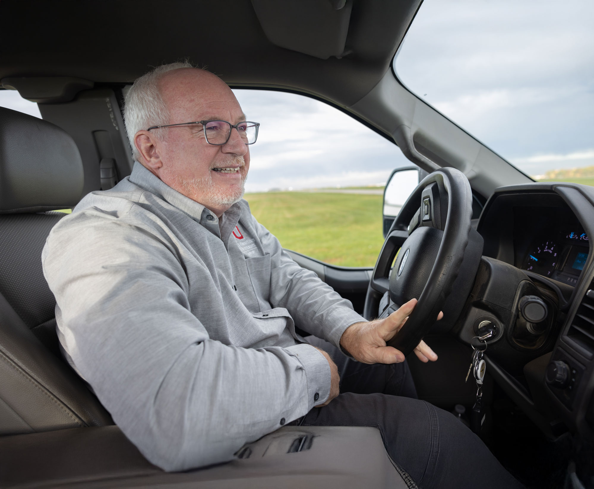 A white man smiles as he sits in the driver’s seat of a truck, with one hand steering the wheel. He’s driving at Ohio State University Airport. Outside his window, there is grassy open space and sky. He has white hair, glasses and a button-up shirt stitch with a block O