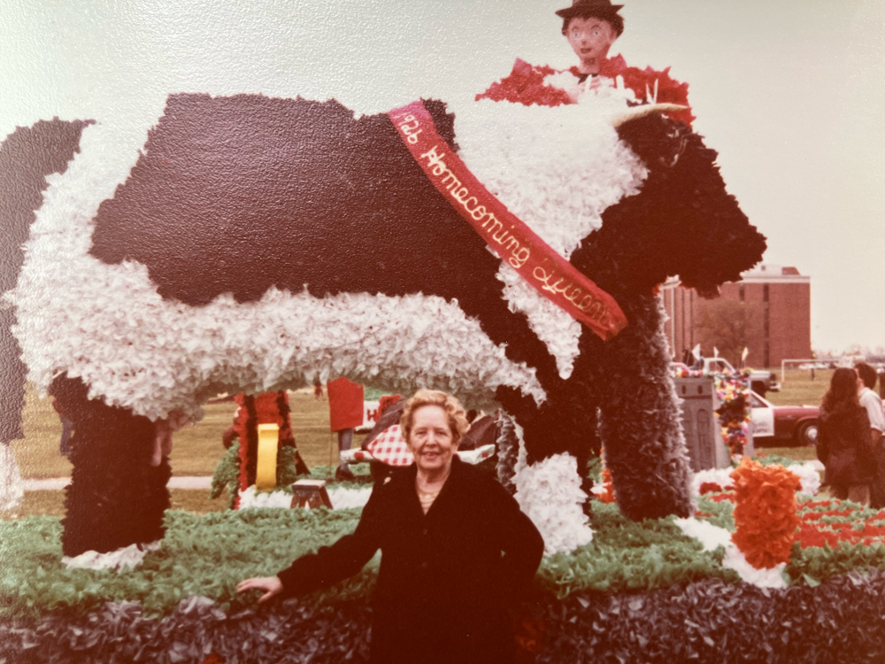 As an older lady, Strapp stands in front of a parade float whith a giant cow wearing a homecoming sash.