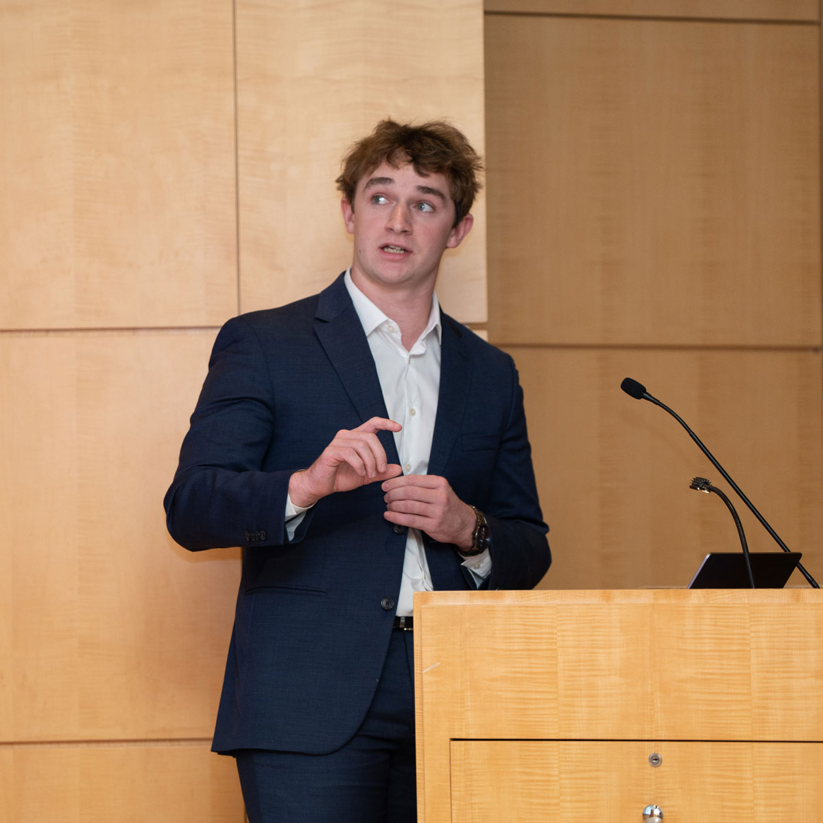 A same young man wears a suit and stands at a lectern, gesturing with one hand while speaking to a research symposium.