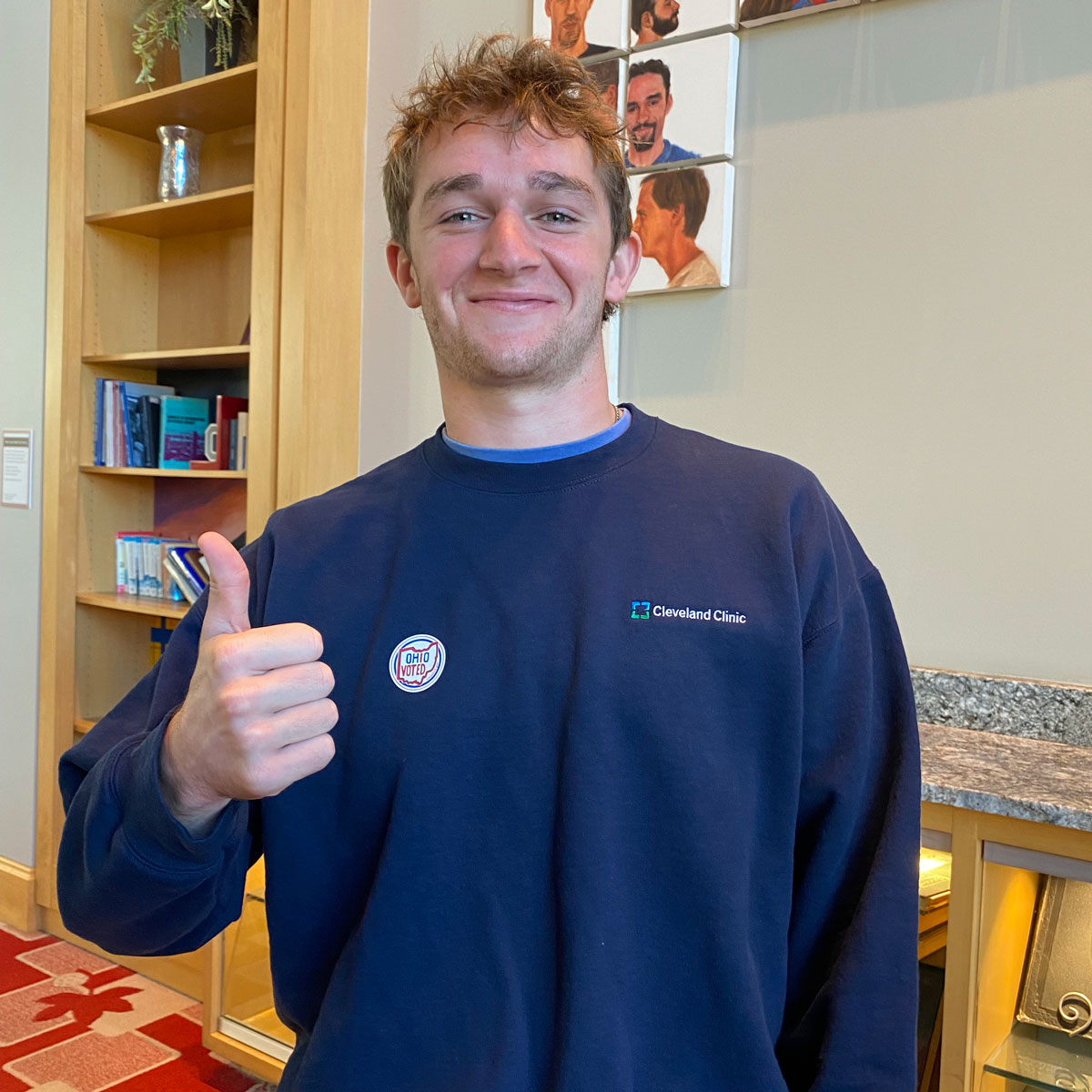 A young man grins with closed lips and messy hair as he gives a thumbs-up inside the Cleveland Clinic. He wears a sweatshirt with a Cleveland Clinic logo and stands near bookshelves and wall art showing illustrated people’s faces.
