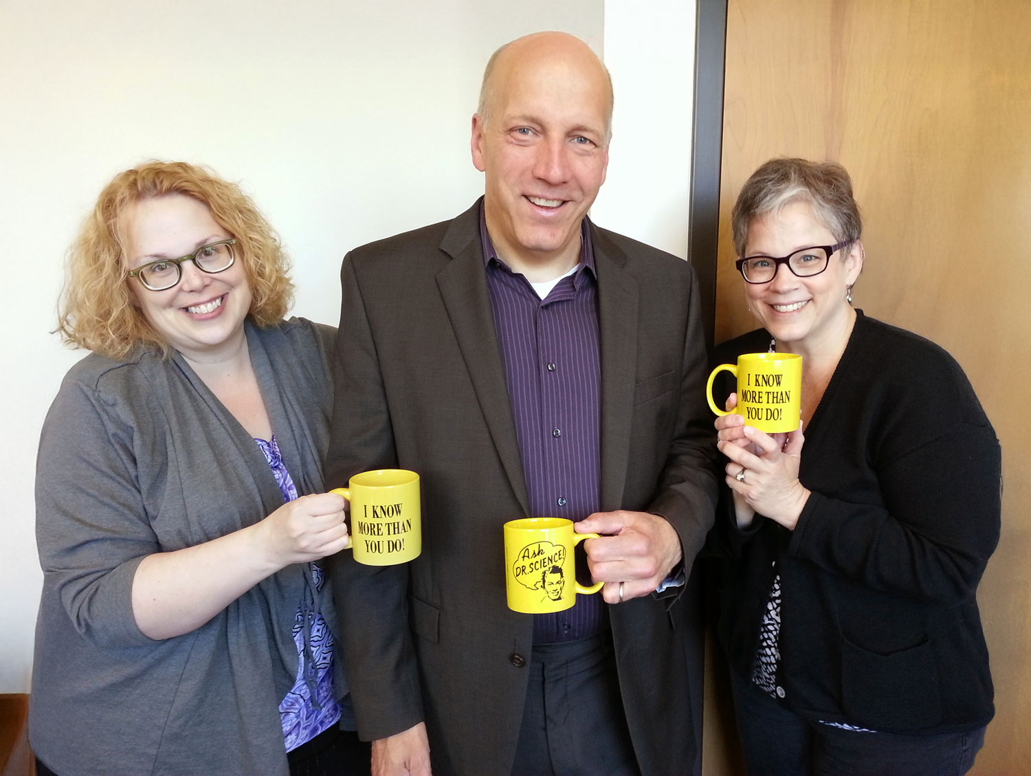 Three Ohio State colleagues smile as they group together to pose for the photo, each holding a yellow mug with printed text. Their closeness speaks to their comfort with each other. The man in the center wears a suit, while a woman stands on both sides of him. They wear layers of business-casual clothes and stand in front of a plain wall and door.
