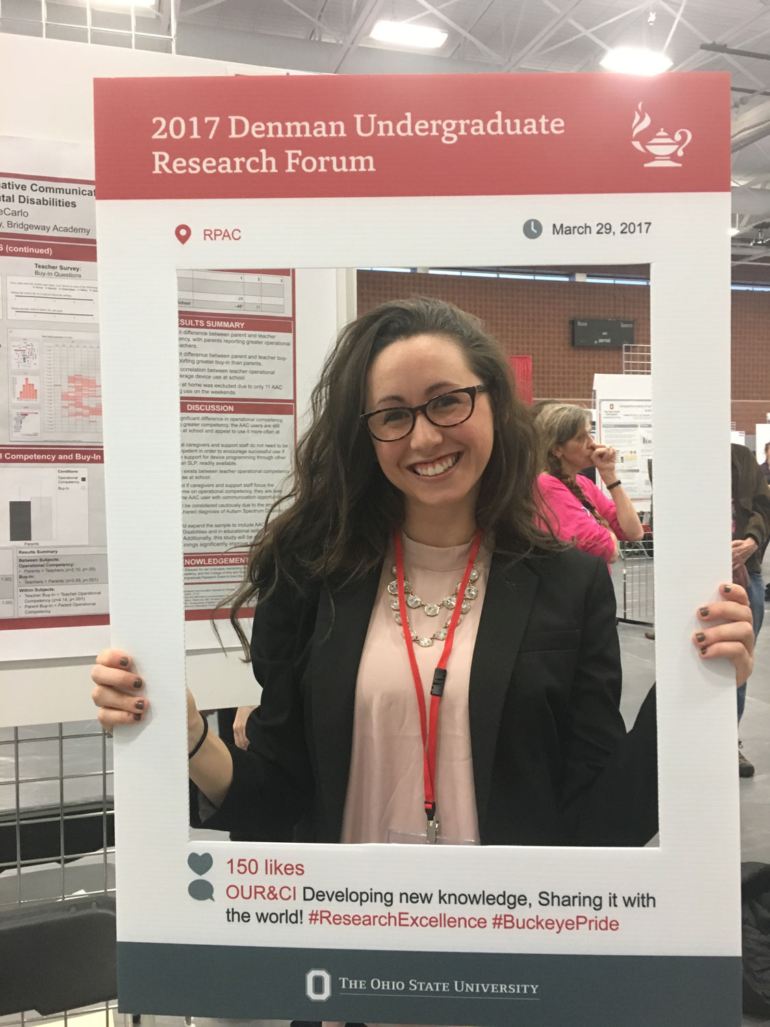A person stands indoors at a research forum, holding a large photo‑frame prop labeled “2017 Denman Undergraduate Research Forum.” The person has long hair, is wearing a jacket over a top with layered necklaces, and has a lanyard around the neck. Research posters and several other people are visible in the background.