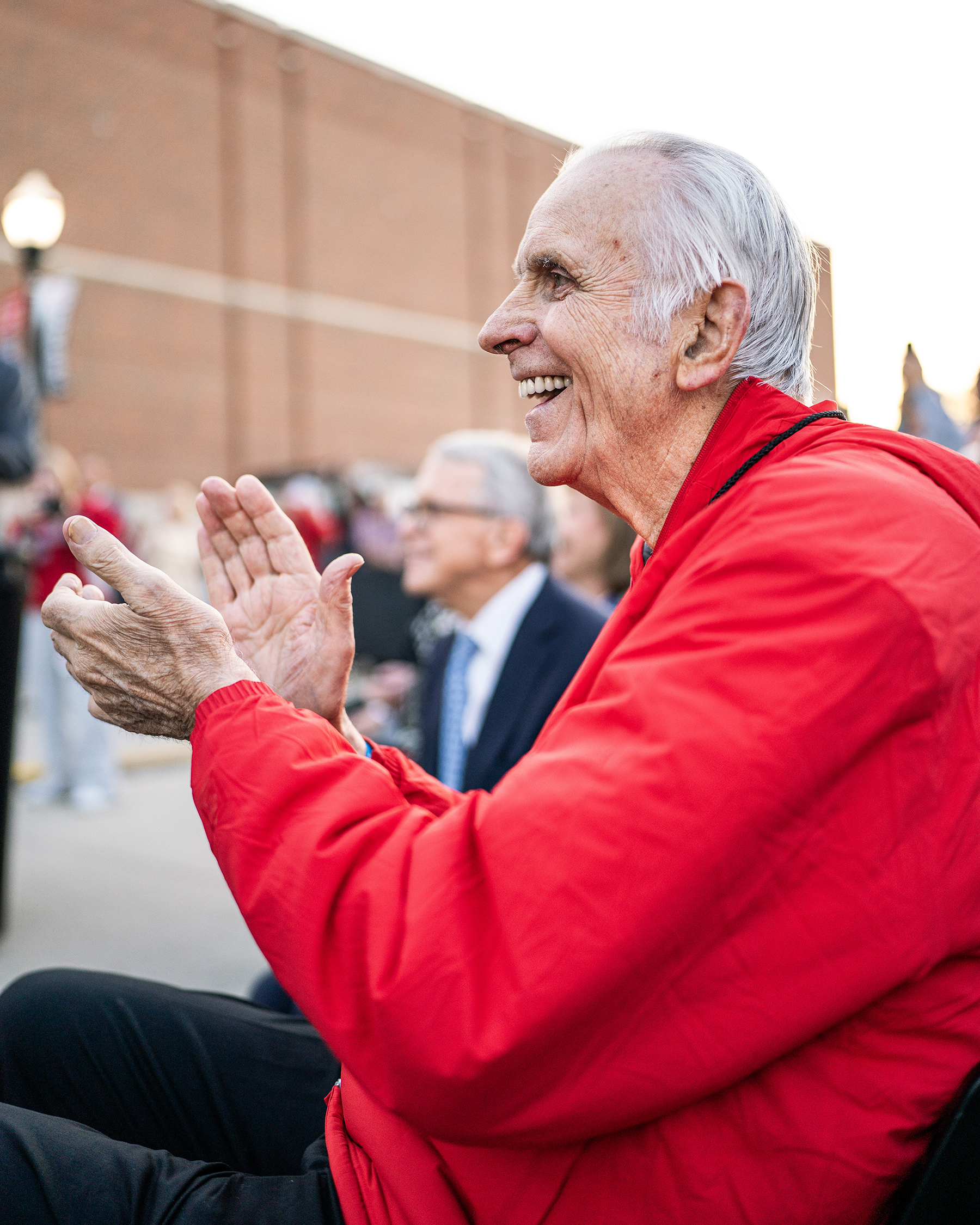 In a photo from the statue's unveiling, Jerry Lucas grins and claps as the cover is pulled off the statue . He wears a scarlet jacket and his face shows smile lines developed over years of his life. He seems happy and proud.