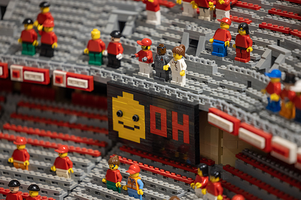 Lego people stand as fans within the model stadium. They include a black man in a business suit and traditionally yellow characters such as construction workers and a doctor. A scoreboard says “O H.”