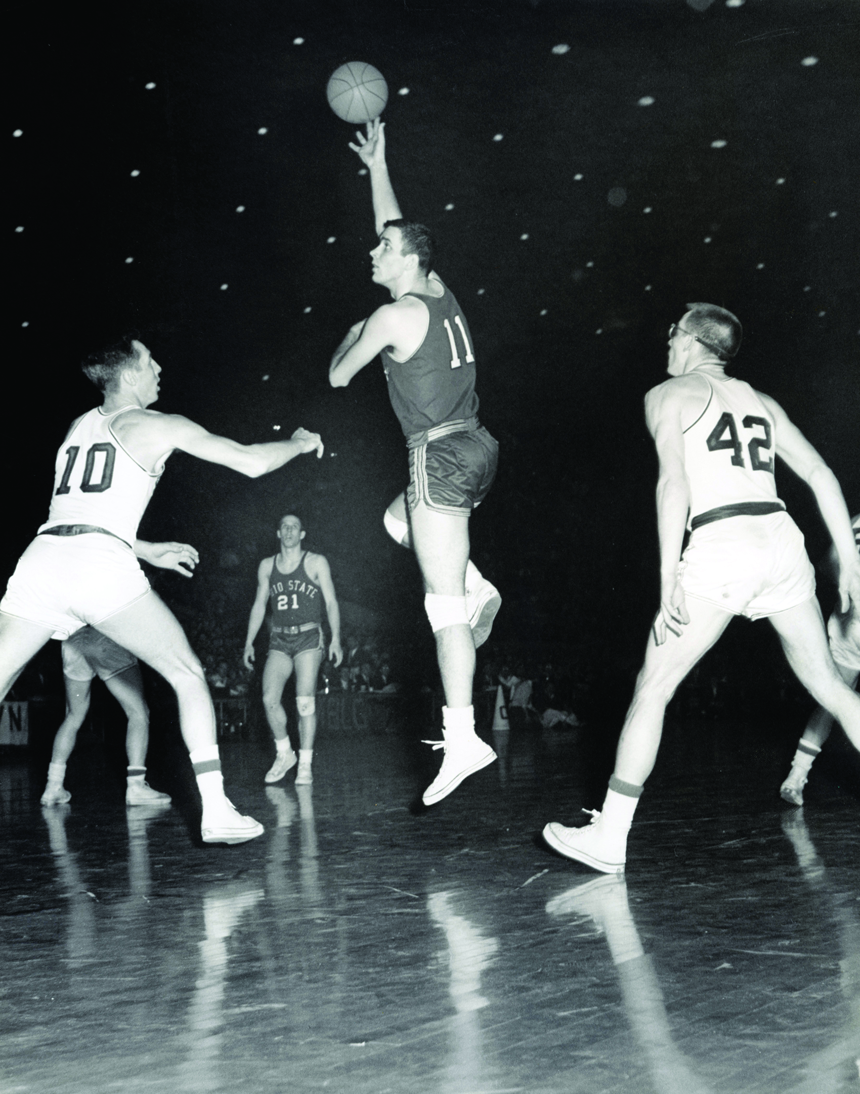 In an older black and white photo, an Ohio STate basketball olayer (Jerry Lucas) jumps high off the floor of the basketball court and hooks the ball over his head. Players from other teams look mini as he rises so high above them. His jersey is number 11 and he's. ayoung white man with neatly shorn hair and a strong build. He also wears knee pads.