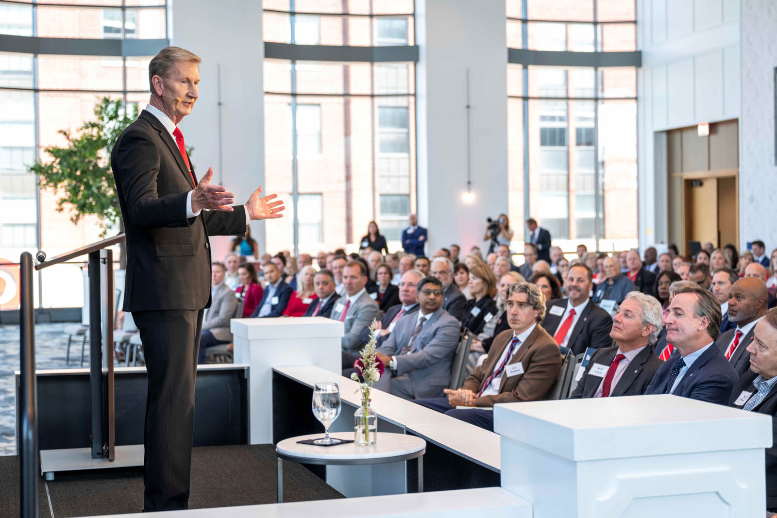 Ohio State President Ted Carter gestures as he speaks to an audience in a bright event space with tall windows. Attendees sit in rows facing him, listening with rapt attention. They’re all wearing suits or similar formal attire. 