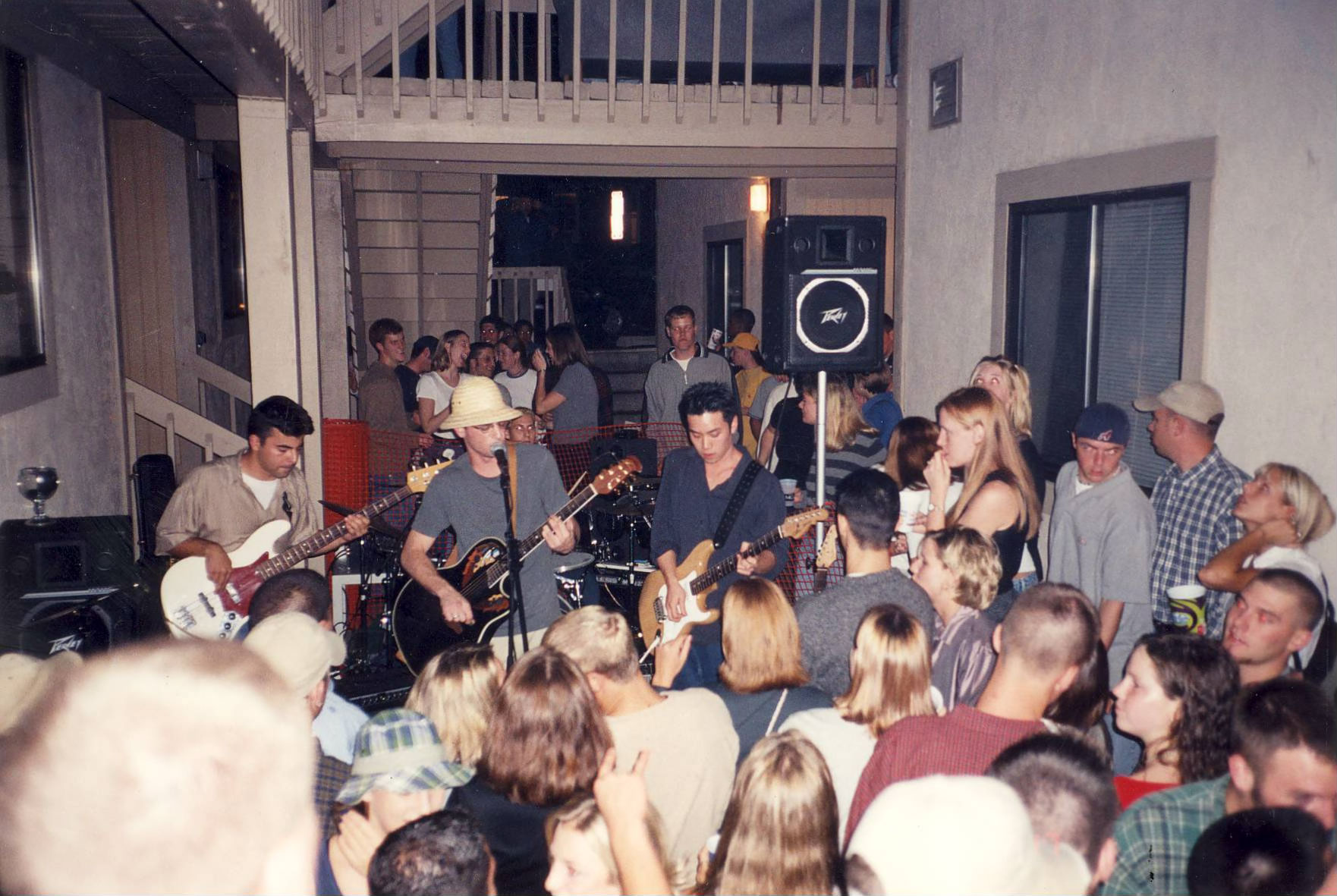 A band performs in a crowded apartment courtyard. The musicians play guitars, bass and drums at the center of the scene, surrounded closely by a large audience. The space has upper walkways, railings, and doors visible in the background, with people gathered on both levels to watch the performance.