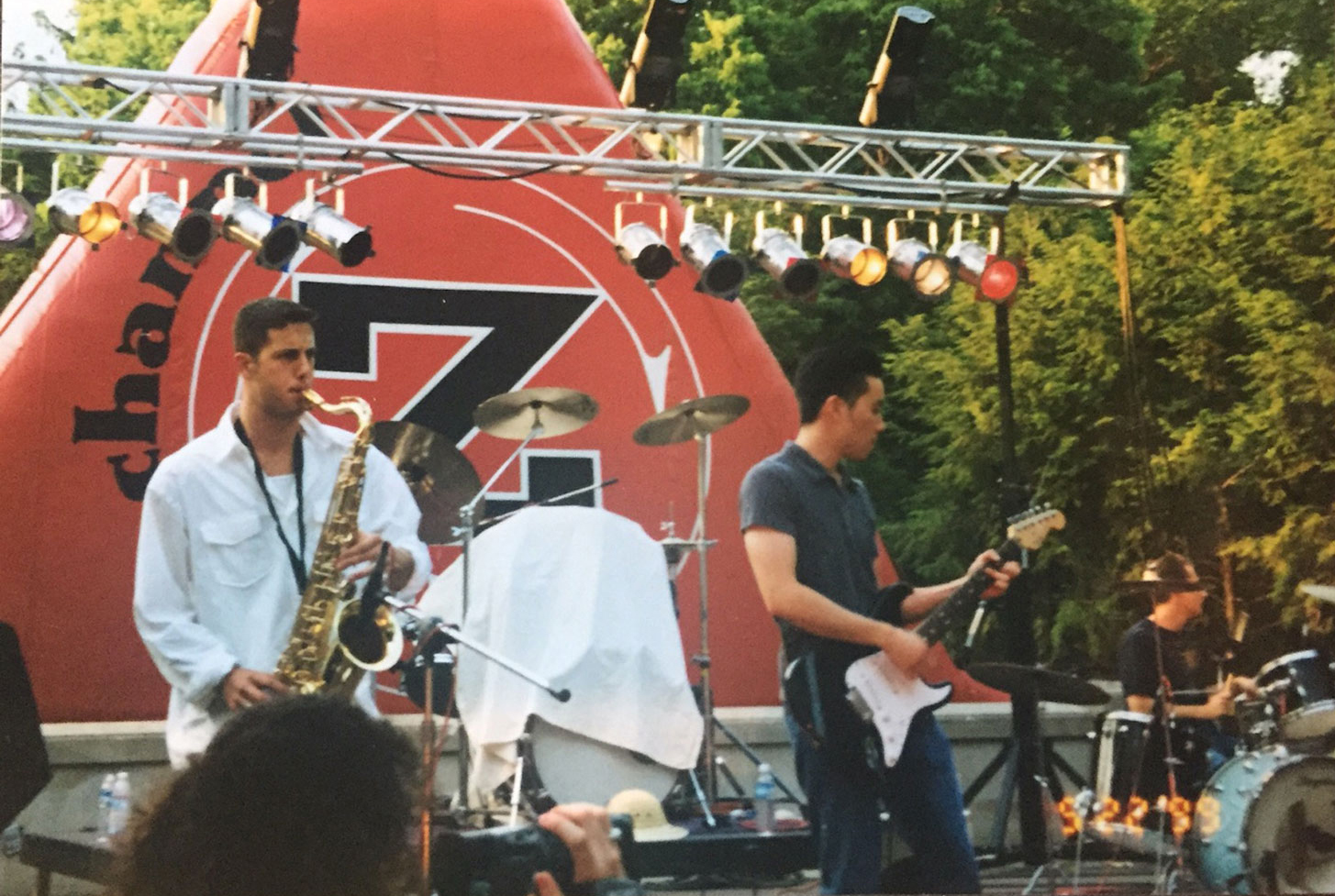 At a small Columbus festival, several musicians perform on an outdoor stage. One plays a saxophone, another plays an electric guitar, and a third plays a drum set. Trees fill the background.