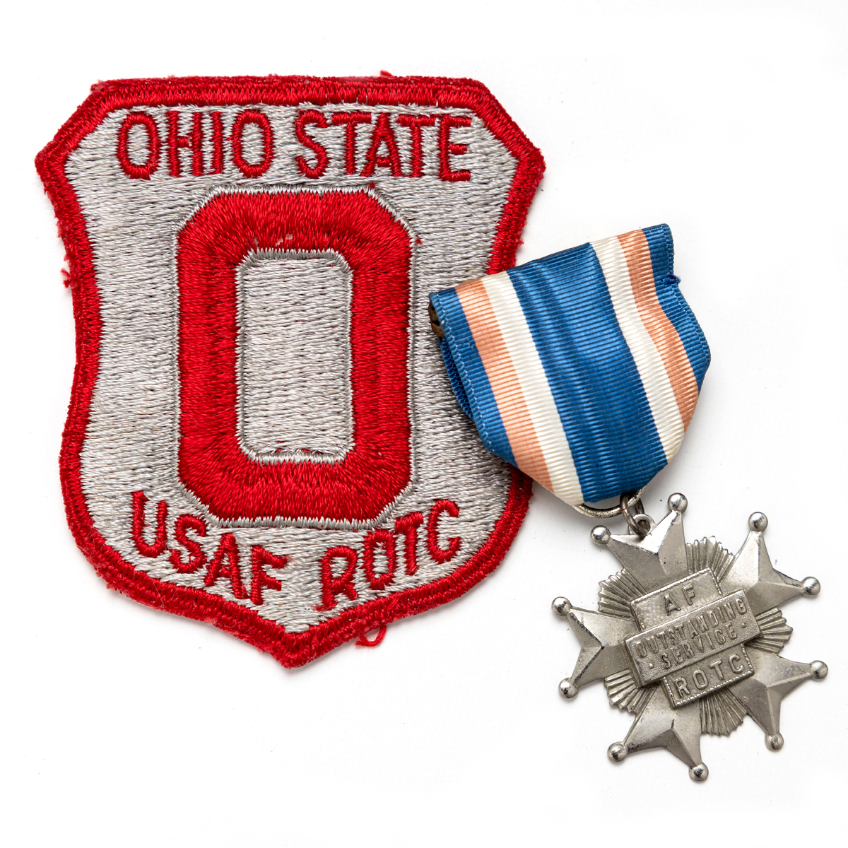 A patch and medal slightly overlap. The scarlet and gray patch says Ohio State USAF ROTC and features a block O. You can see the embroidery threads used to make it. The star-shaped medal hangs from a blue, peach and white ribbon. The medal says AF ROTC Outstanding service.