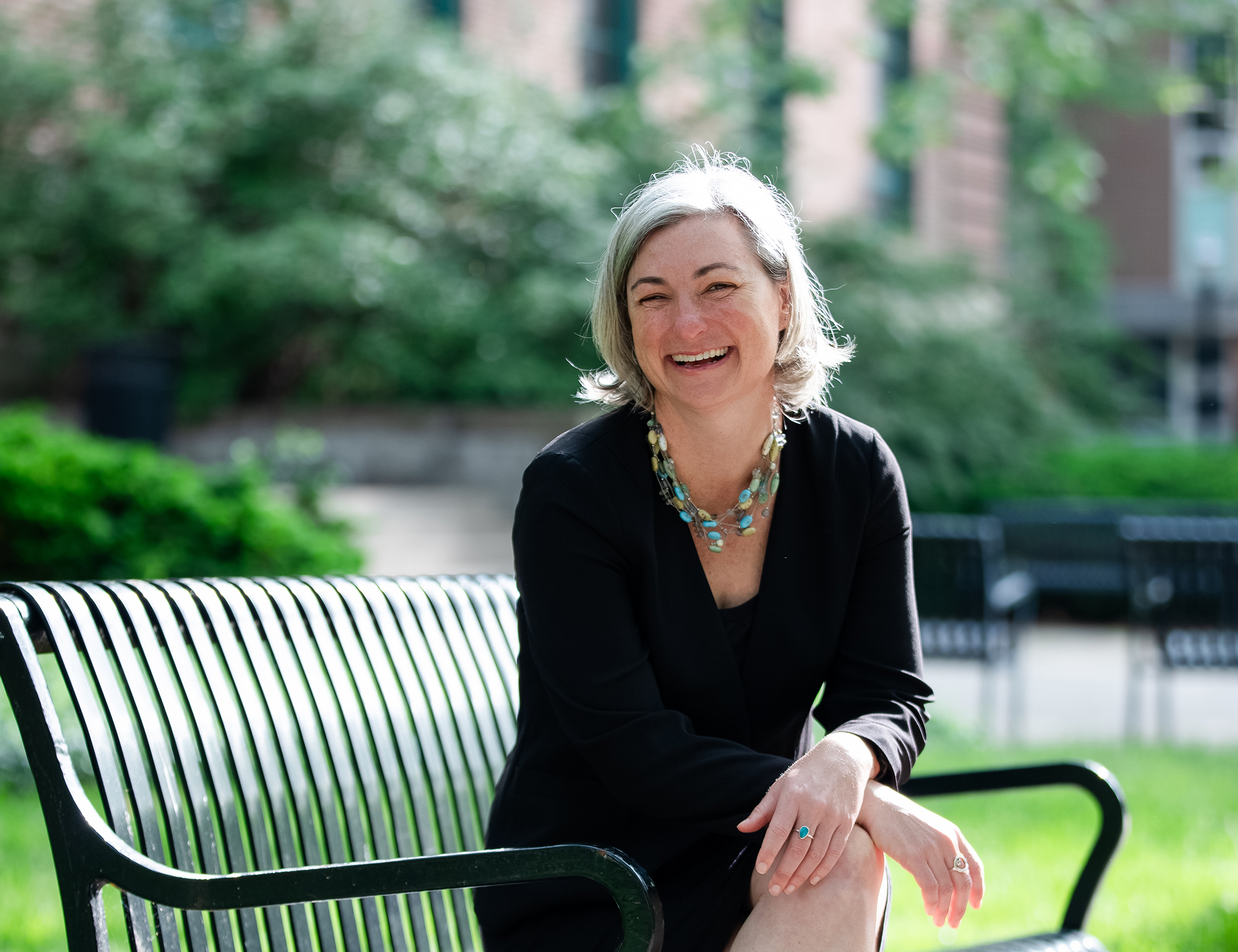 A white woman with shoulder-length gray hair but a young face laughs as she is photographed. Her enjoyment is so true, her eyes are completely squinted. To be fair, it is a bright day on campus. She sits on a metal bench.