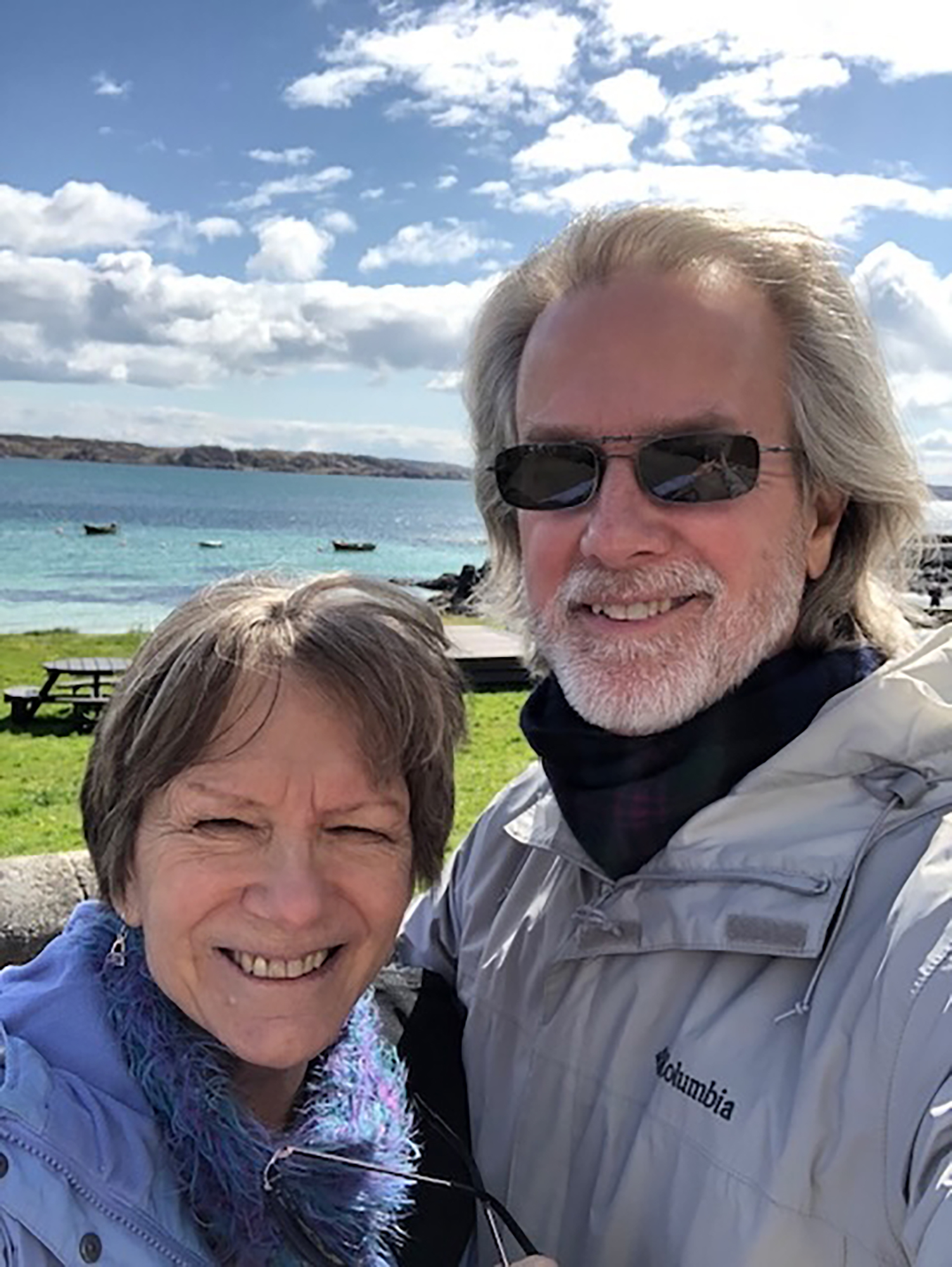 Roy and June smile as they stand close on a seashore on a very bright day in Ireland. They’re dressed in hiking jackets and look happy.