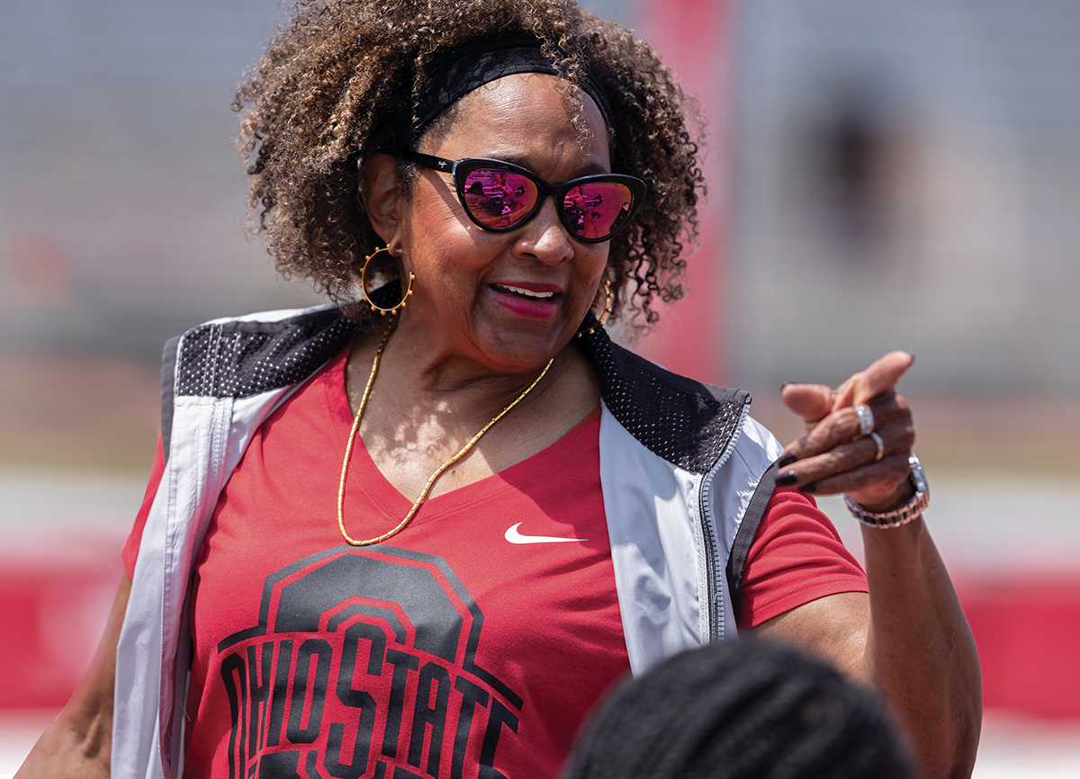 An older black woman wearing an Ohio State T-shirt, mirrored sunglasses, gold jewelry and a smile points while coaching outside.