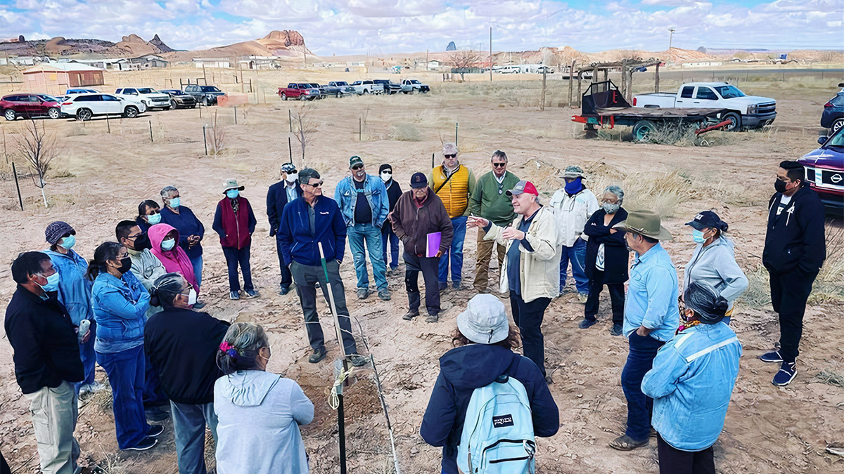 Two dozen people stand in a circle listening to two male teachers in a dry, dirt field.