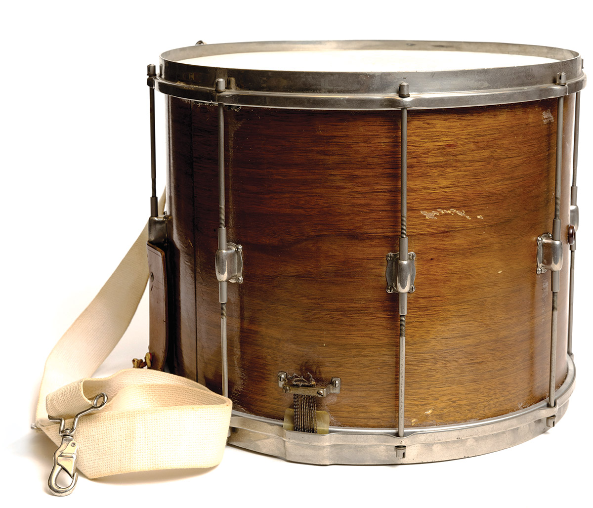 An old snare drum has wooden shell stained a dark golden brown, a white drum head and silver-colored rims and wires connecting top to bottom. An off-white strap shows how the drummer carried it.