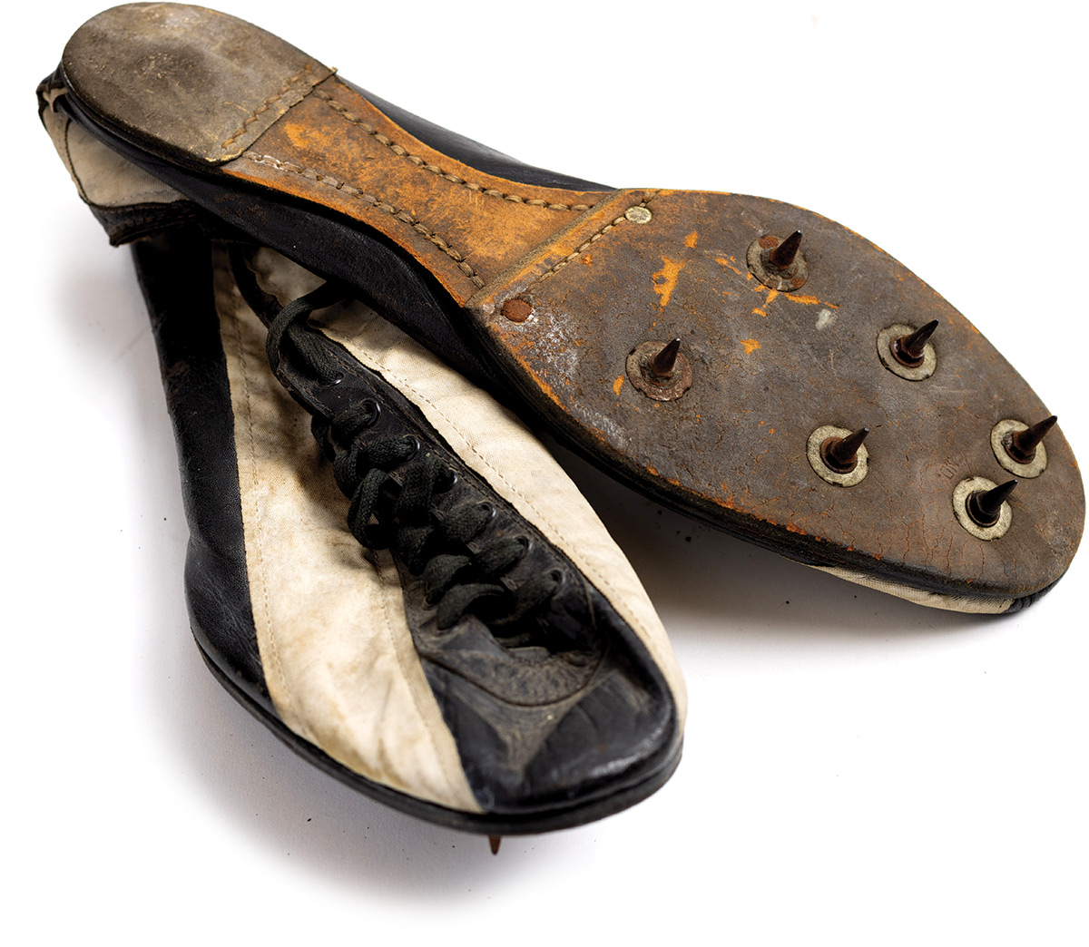 A pair of worn track shoes are black and white lace-ups. One is turned upside down to show the blackened sole and six thin, black spikes, all on the portion of the shoe that would support the toes and ball of the foot.